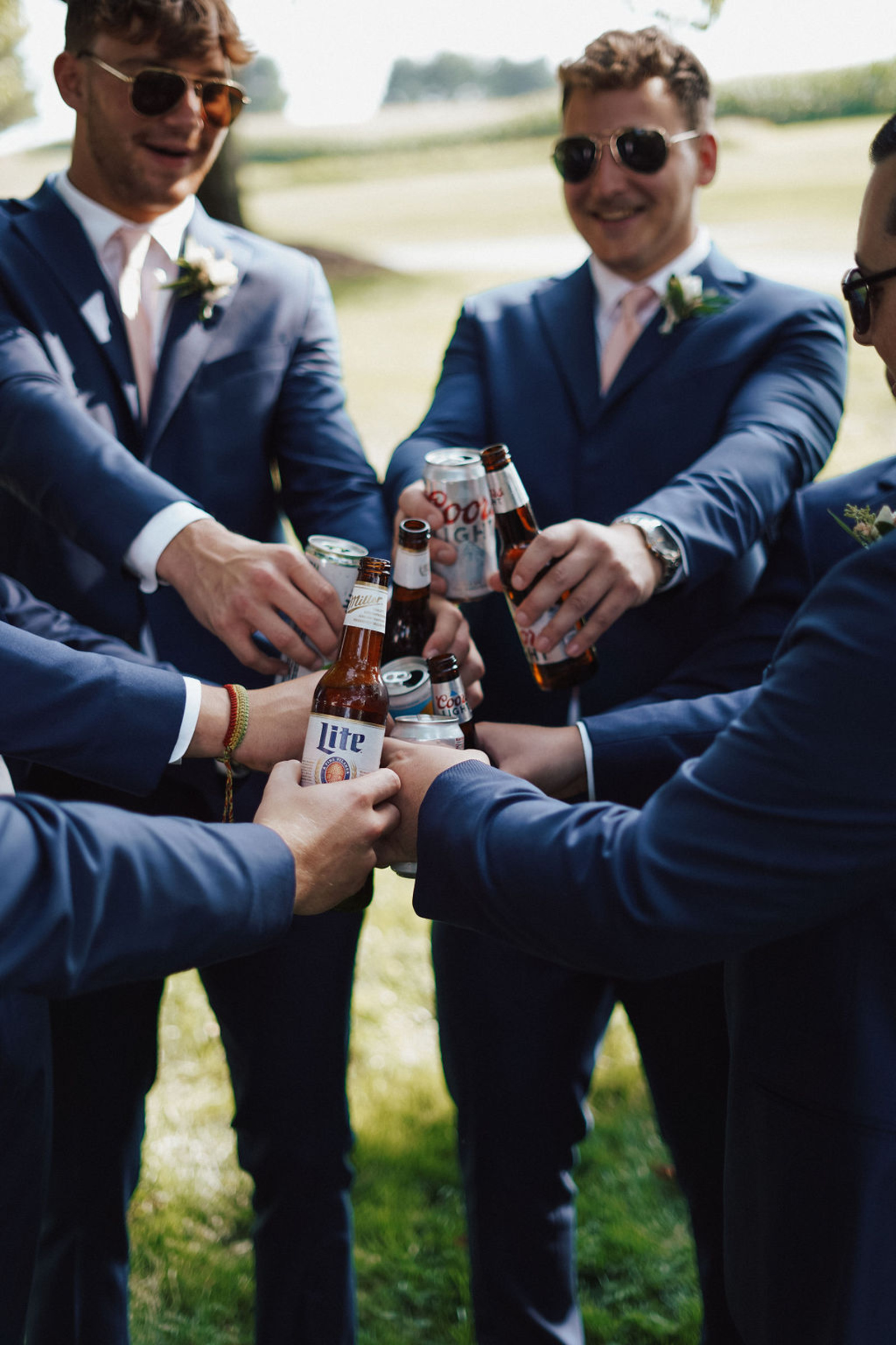 Central PA groom and groomsmen in navy suits standing in a circle clinking beers together to say "cheers"