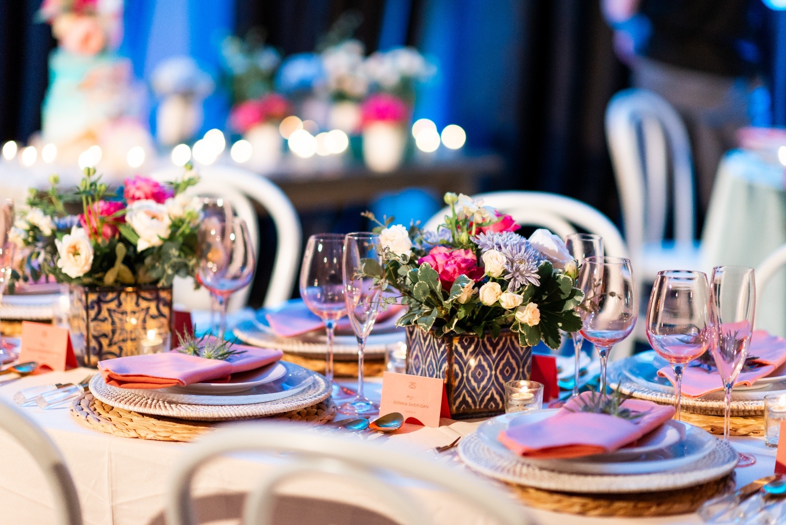 Elegantly set table with white chairs, colorful floral centerpieces, pink napkins, and glowing candles, creating a festive and inviting ambiance.