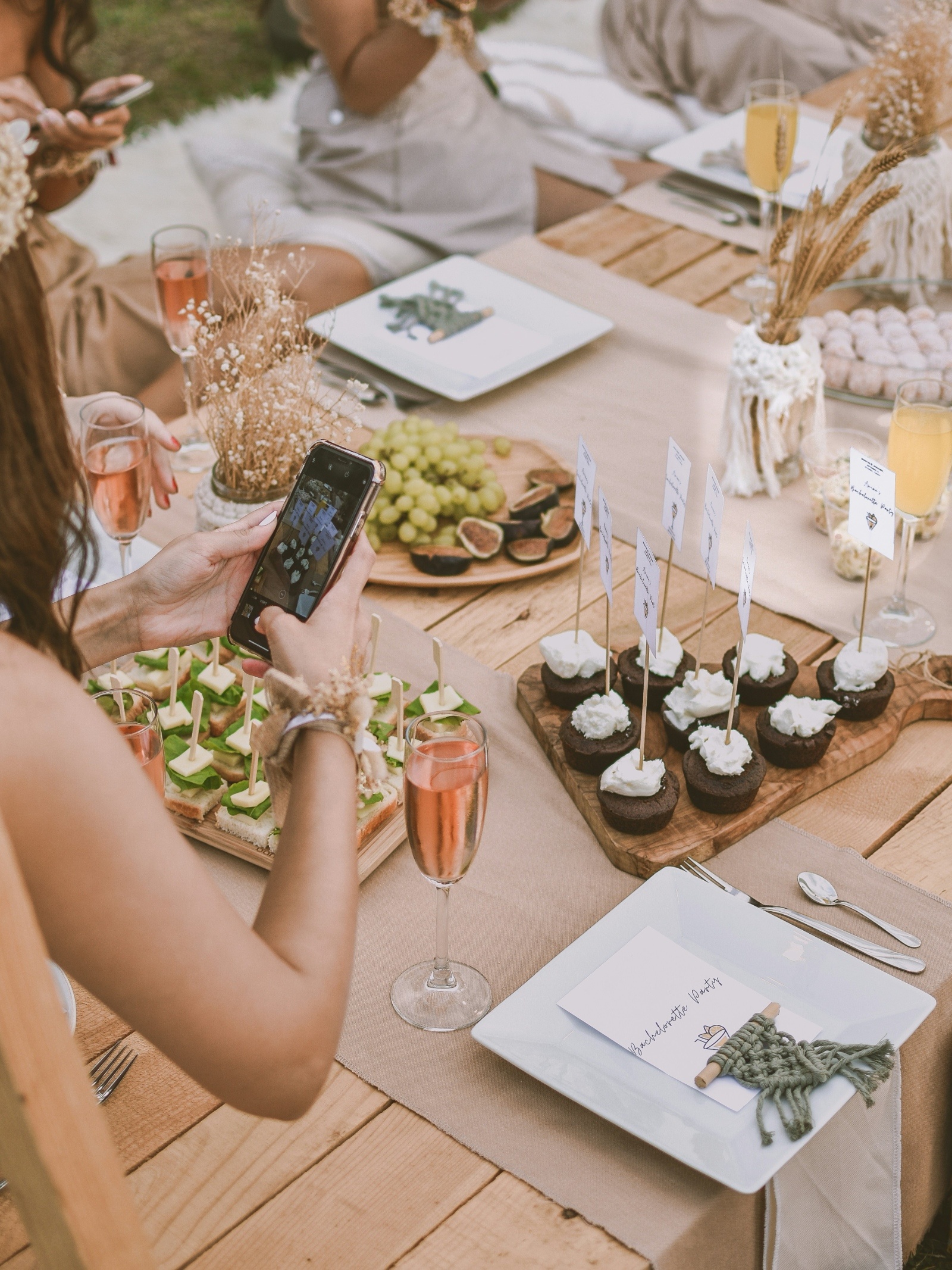 A beautifully styled picnic table with charcuterie, cupcakes, and rosé surrounded by friends capturing photos — reflecting the relaxed, wine-country charm of a Finger Lakes bachelorette weekend.