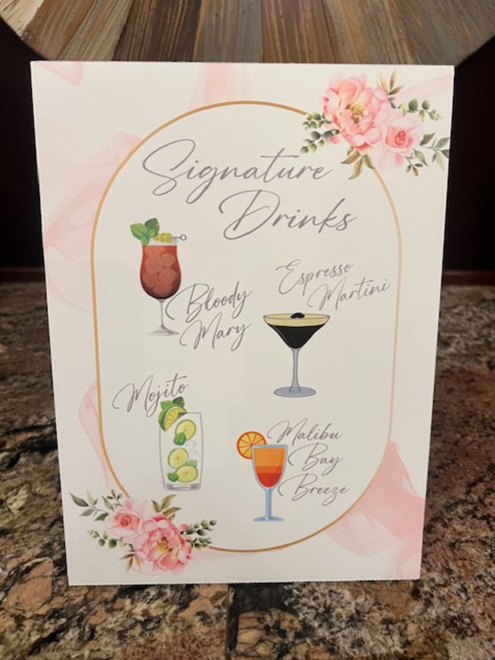 A floral-themed signature drink sign displays illustrated cocktails including a Bloody Mary, Espresso Martini, Mojito, and Malibu Bay Breeze on a granite countertop.