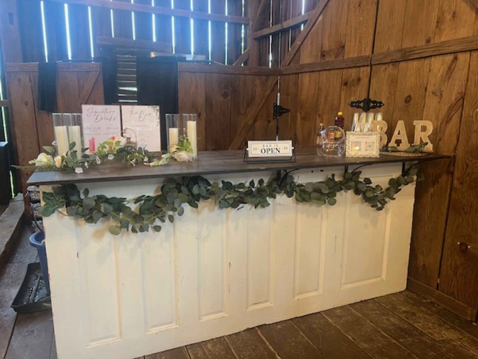 A rustic wedding bar decorated with candles, greenery, and floral accents features a sign reading “Bar is Open” and a signature drink menu against a wooden barn backdrop.