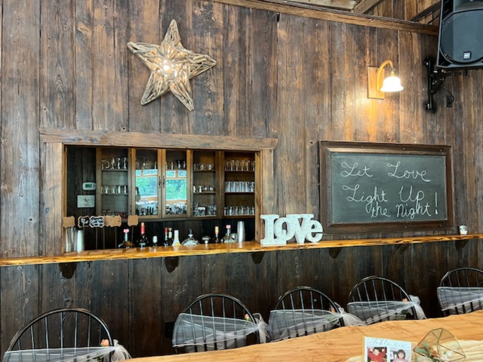 A cozy barn bar with wooden walls, a glowing star light, and a chalkboard sign reading “Let Love Light Up the Night,” accented by glassware and a decorative “LOVE” sign.