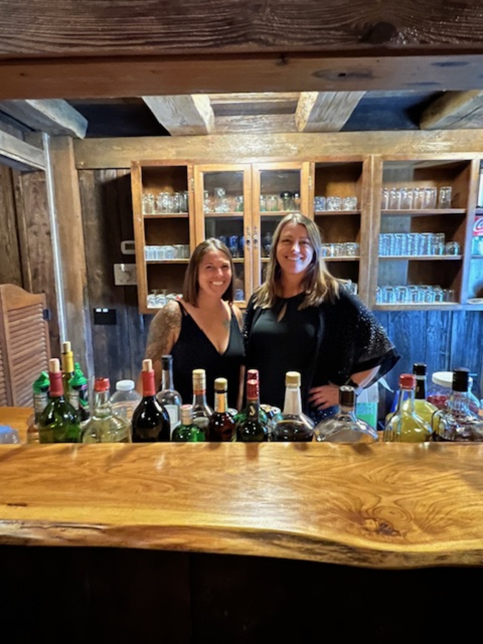 Two smiling bartenders stand behind a rustic wooden bar lined with bottles and glassware, representing The Mixology Masters event bartending team.