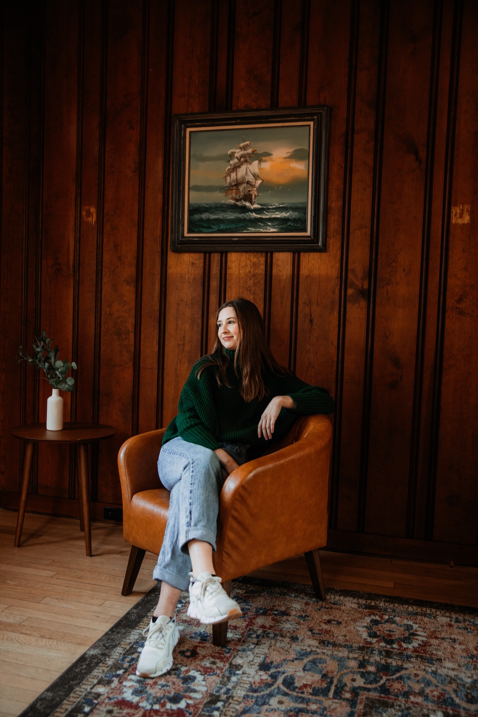 Photographer Téa Ceresini sits in a leather chair by a wooden wall, smiling thoughtfully beneath a nautical painting.