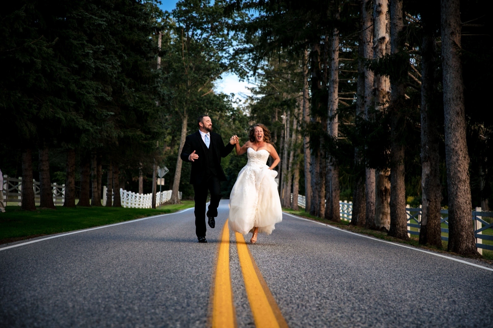 Bride and groom run hand in hand down a tree-lined country road during playful wedding portraits at a Pennsylvania wedding venue.