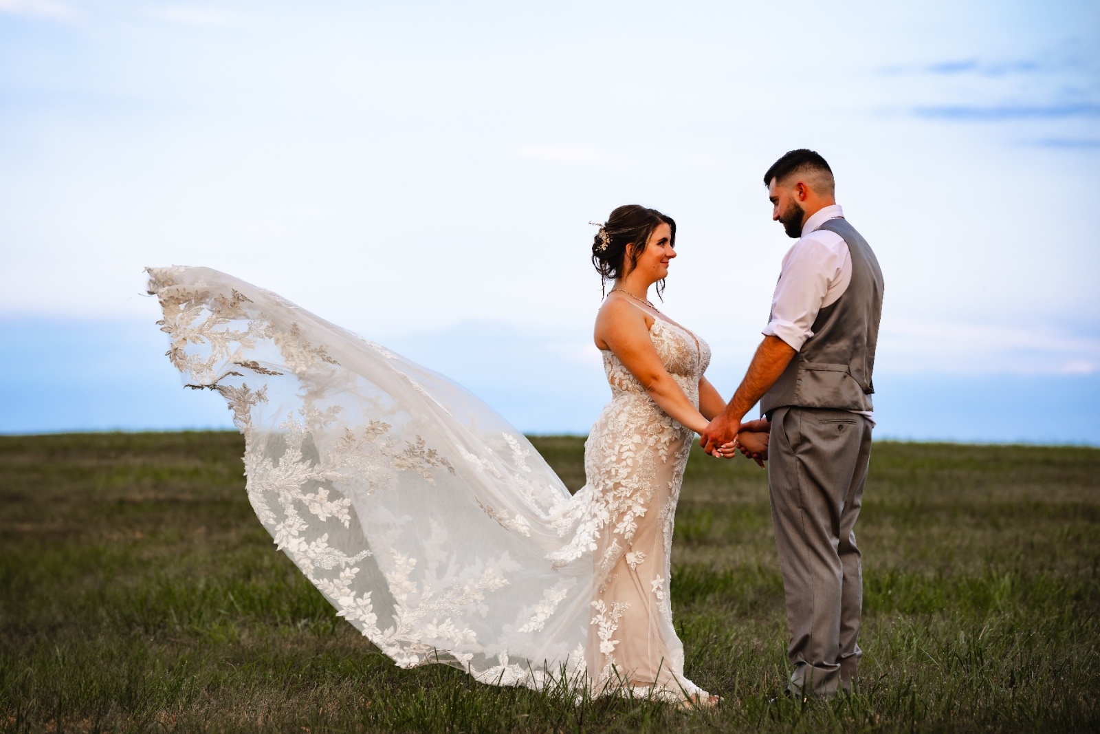 Bride and groom hold hands in an open field as the bride's lace veil flows dramatically in the wind during sunset portraits.