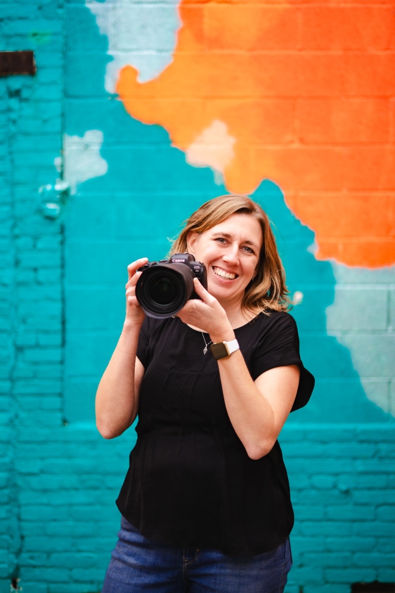 Wedding photographer Kelly Knaub smiles while holding her camera in front of a colorful mural in York, Pennsylvania.