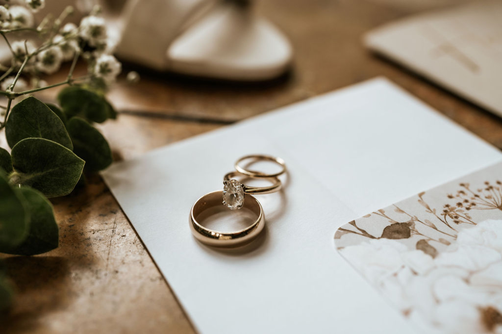 Wedding day details featuring gold rings, vow books, perfume, bridal jewelry, shoes, and a lace clutch arranged beautifully beside a floral invitation for a Moonstone Manor wedding.
