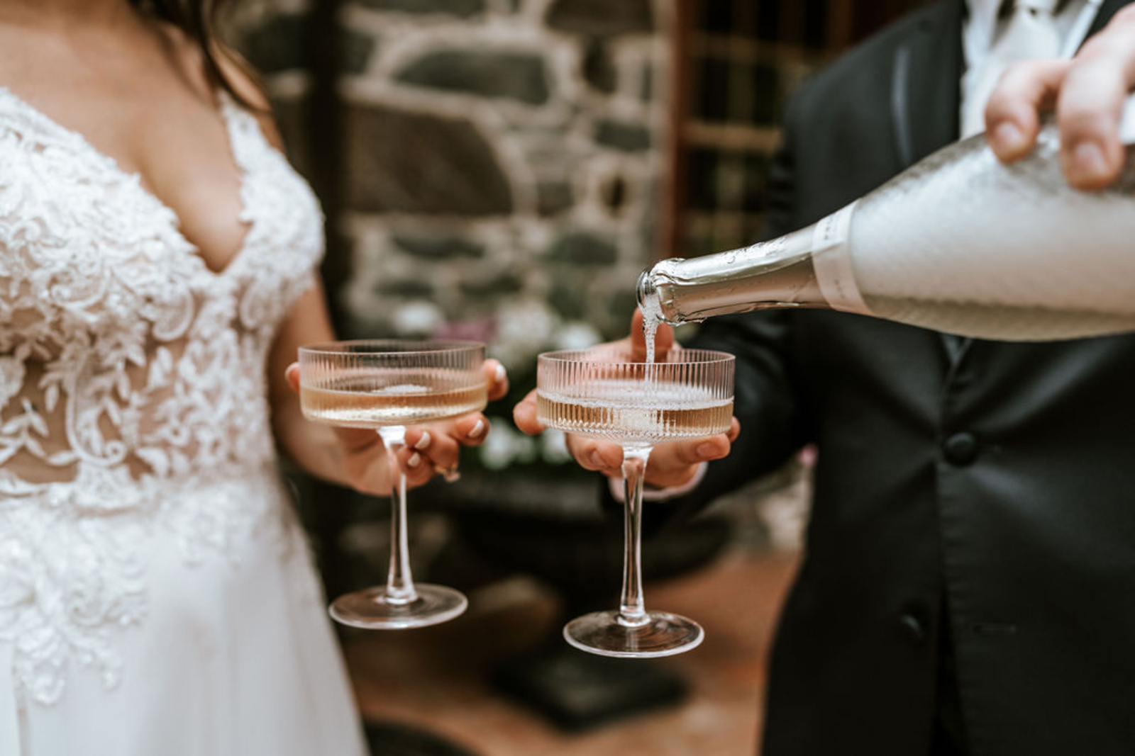 Close-up of the bride and groom pouring champagne into coupe glasses during their wedding celebration at Moonstone Manor.