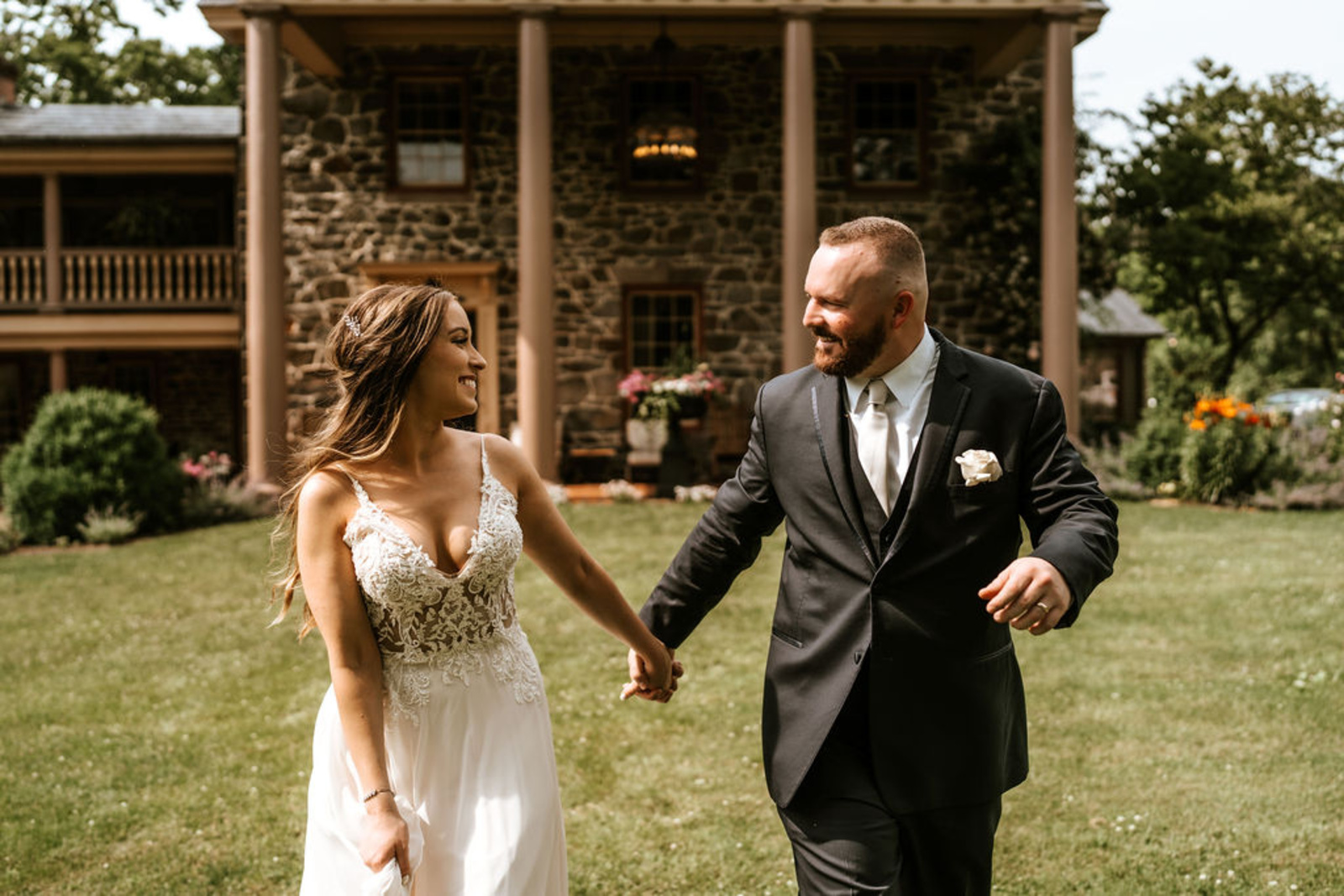 Bride and groom walk hand-in-hand across the manicured lawn in front of the stone mansion at Moonstone Manor, smiling after their ceremony.