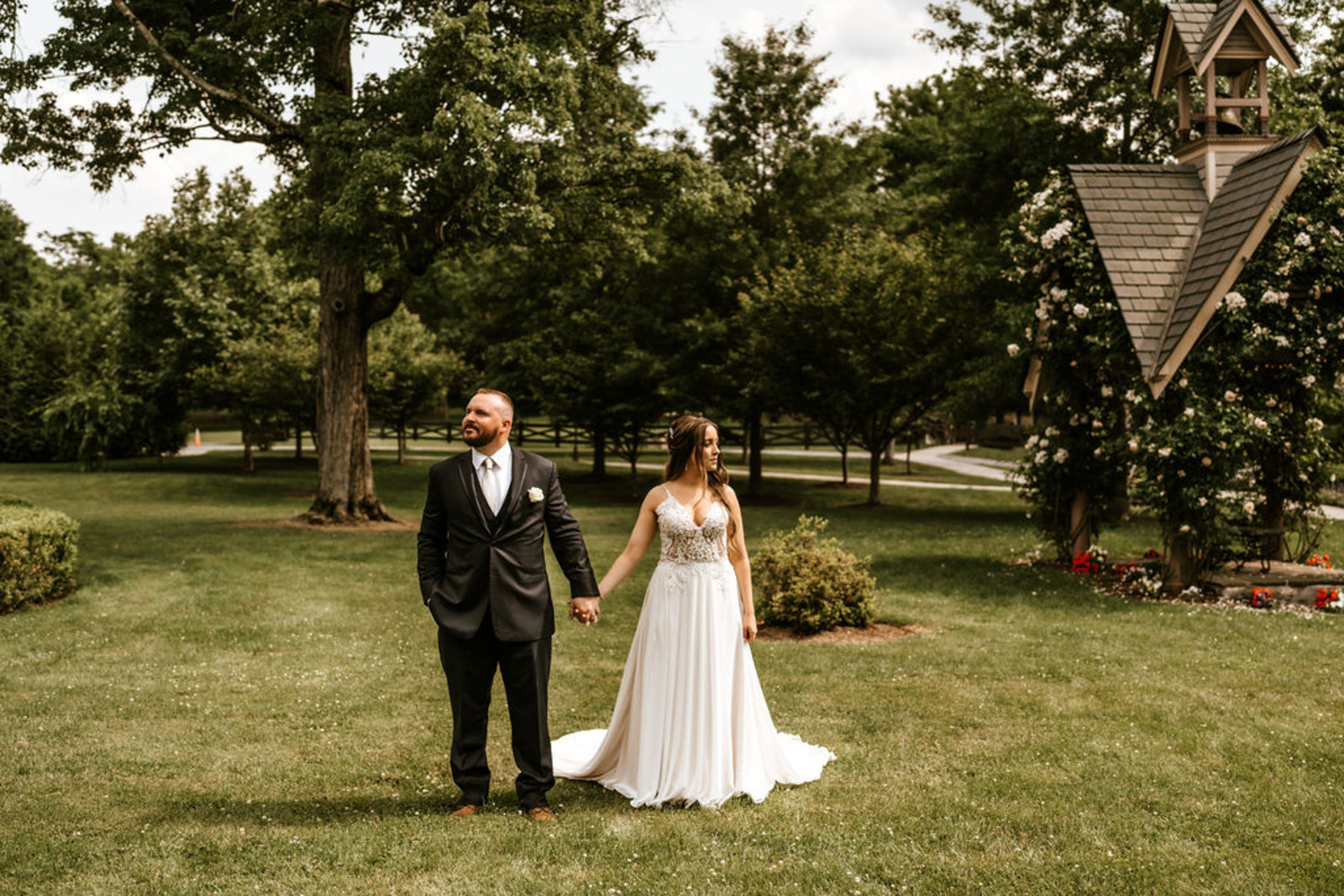 Bride and groom hold hands in front of the chapel garden at Moonstone Manor, surrounded by tall trees and lush summer greenery.