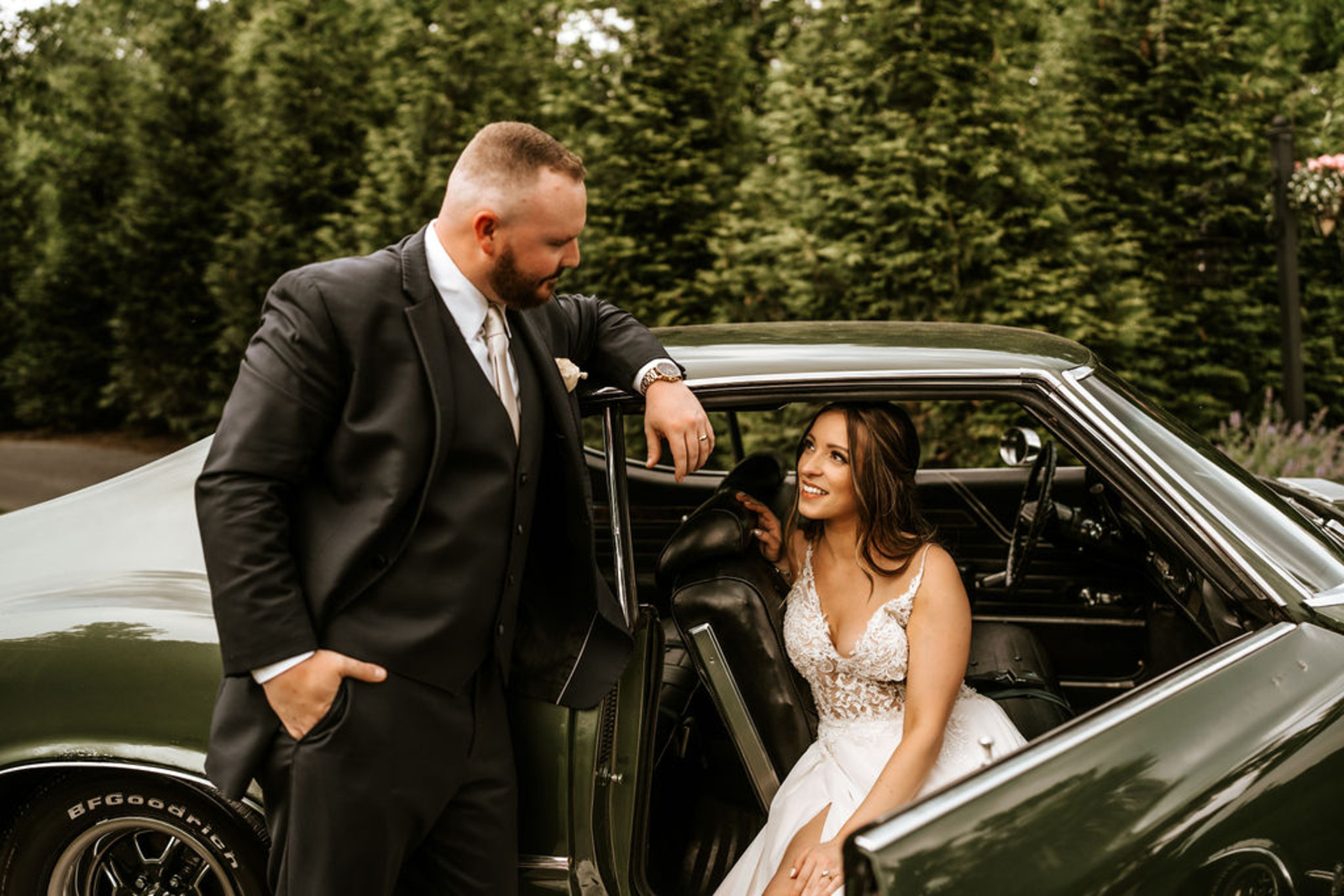Bride smiles up at her groom while sitting in a vintage green car parked outside Moonstone Manor, blending timeless romance and style.