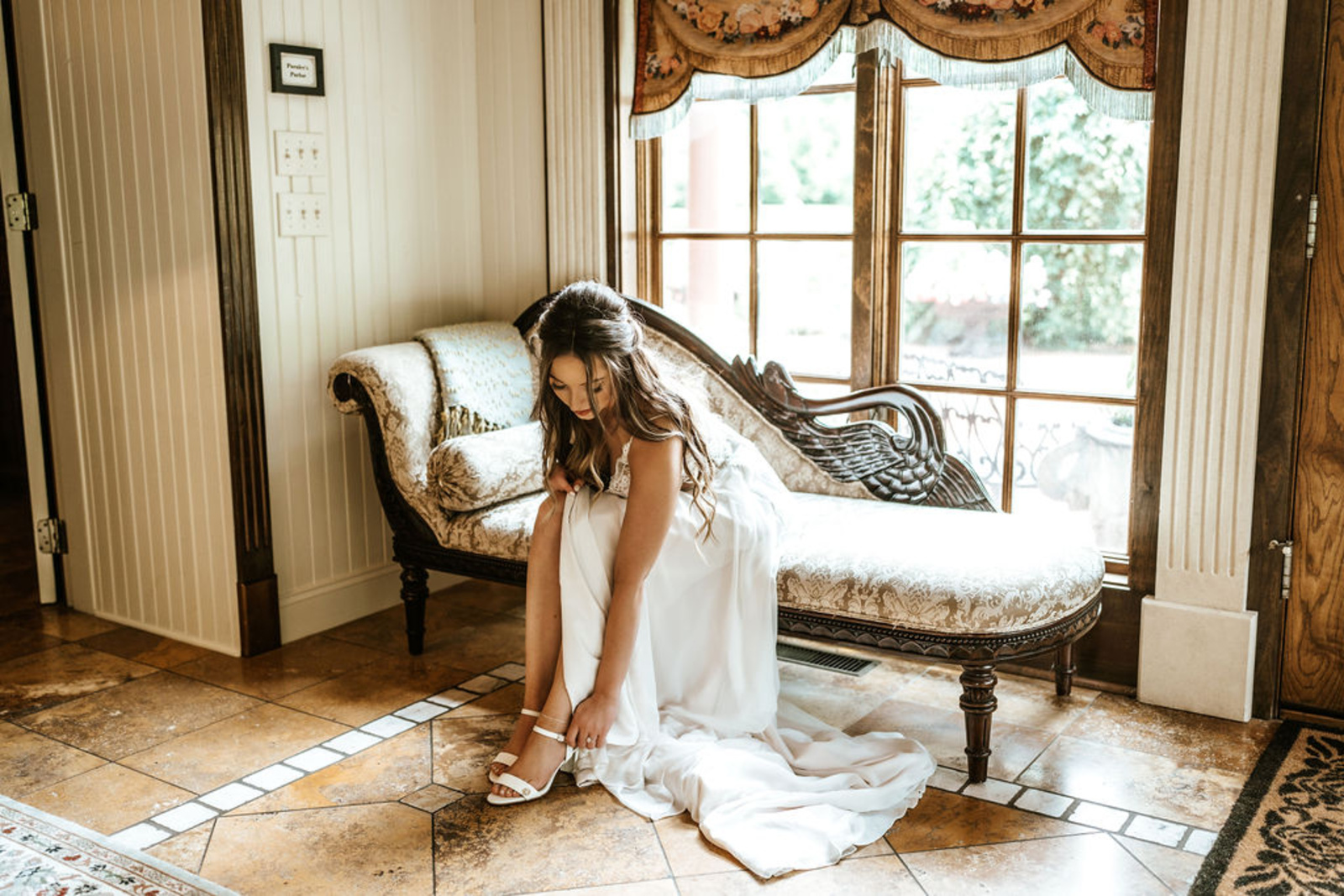 The bride adjusts her white heels while sitting on an elegant vintage chaise inside Moonstone Manor’s bridal suite, surrounded by warm natural light.
