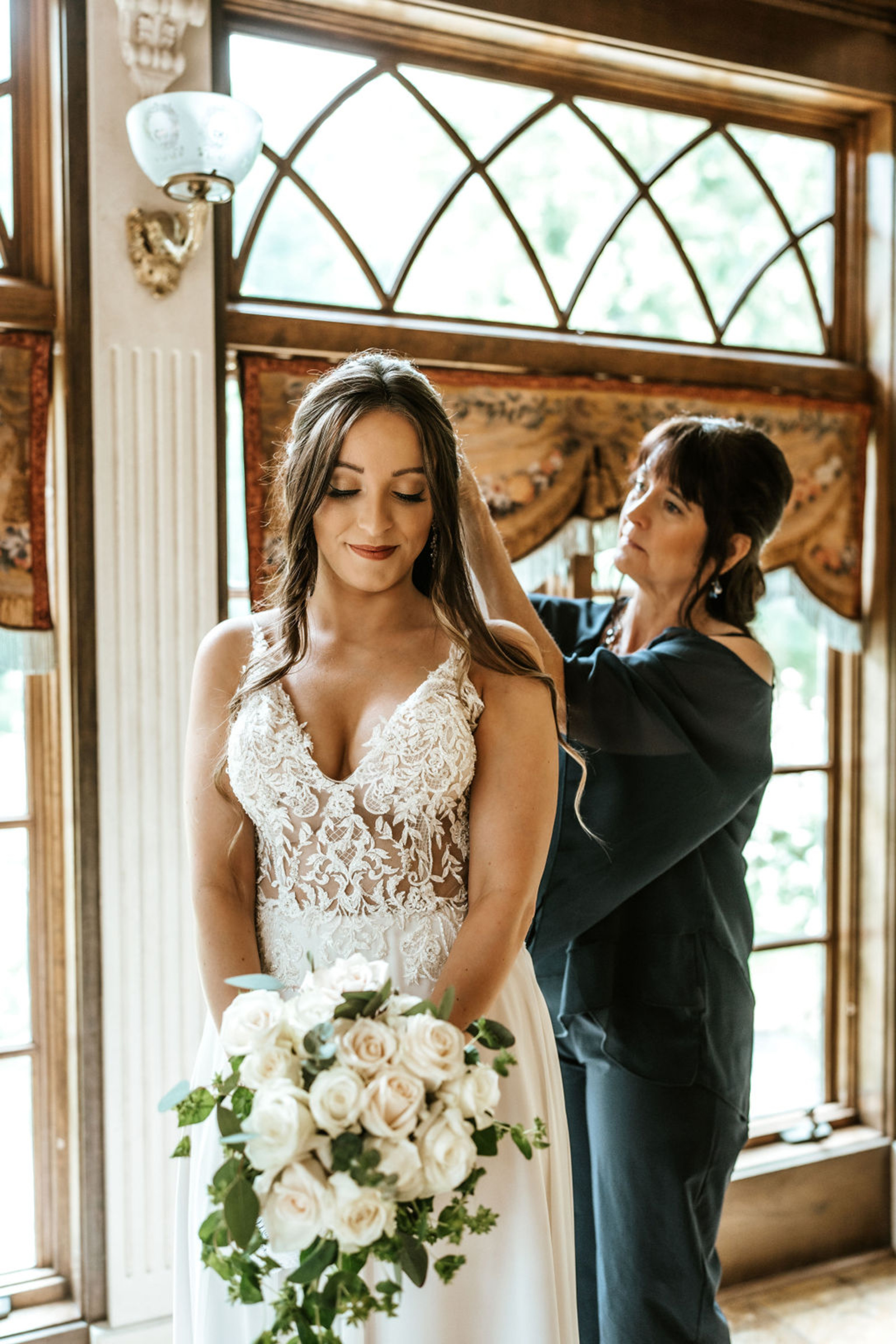 The bride’s mother fastens her daughter’s hair as she holds her bouquet of white roses, sharing a heartfelt getting-ready moment inside Moonstone Manor.