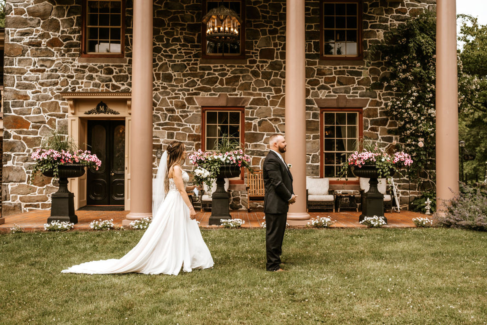 The bride walks toward her groom for their first look in front of Moonstone Manor’s grand stone façade, surrounded by blooming flowers and timeless architecture.