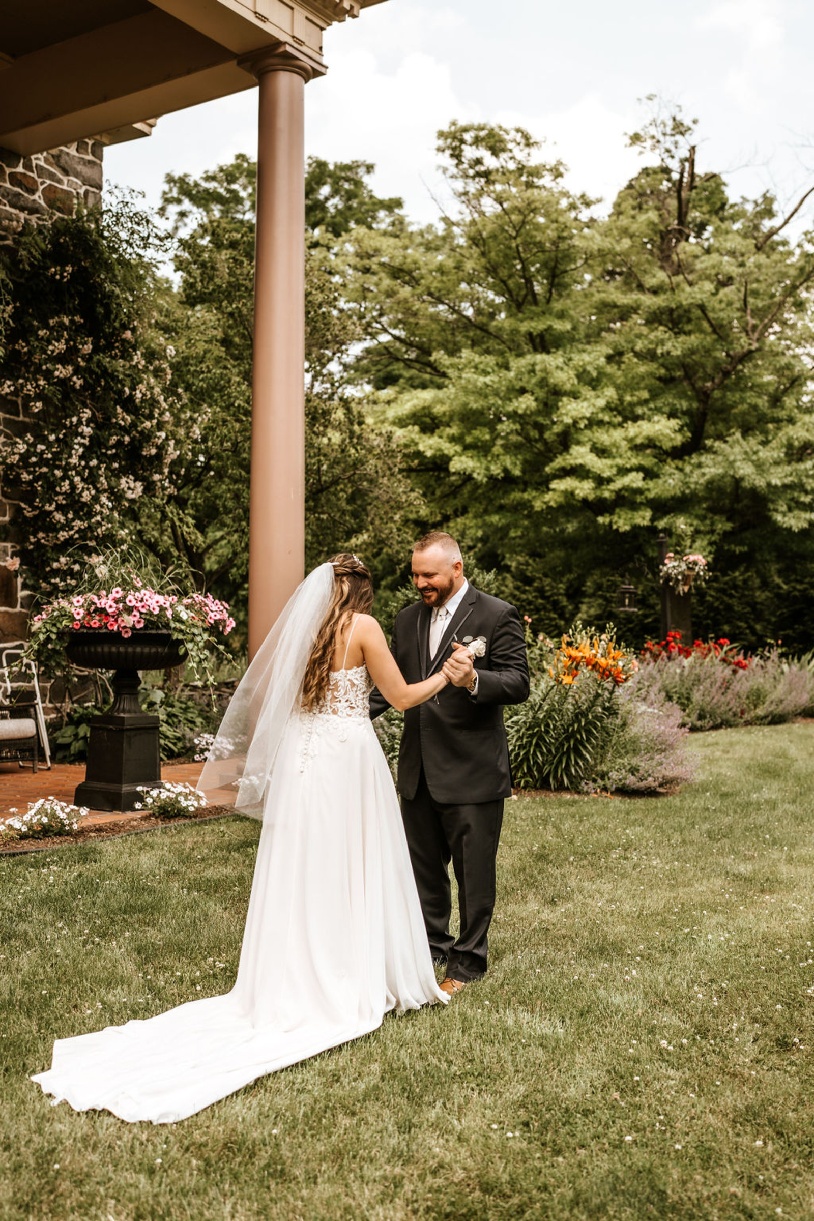 The bride and groom share a joyful first look moment on the lawn of Moonstone Manor, holding hands beneath the venue’s towering columns.