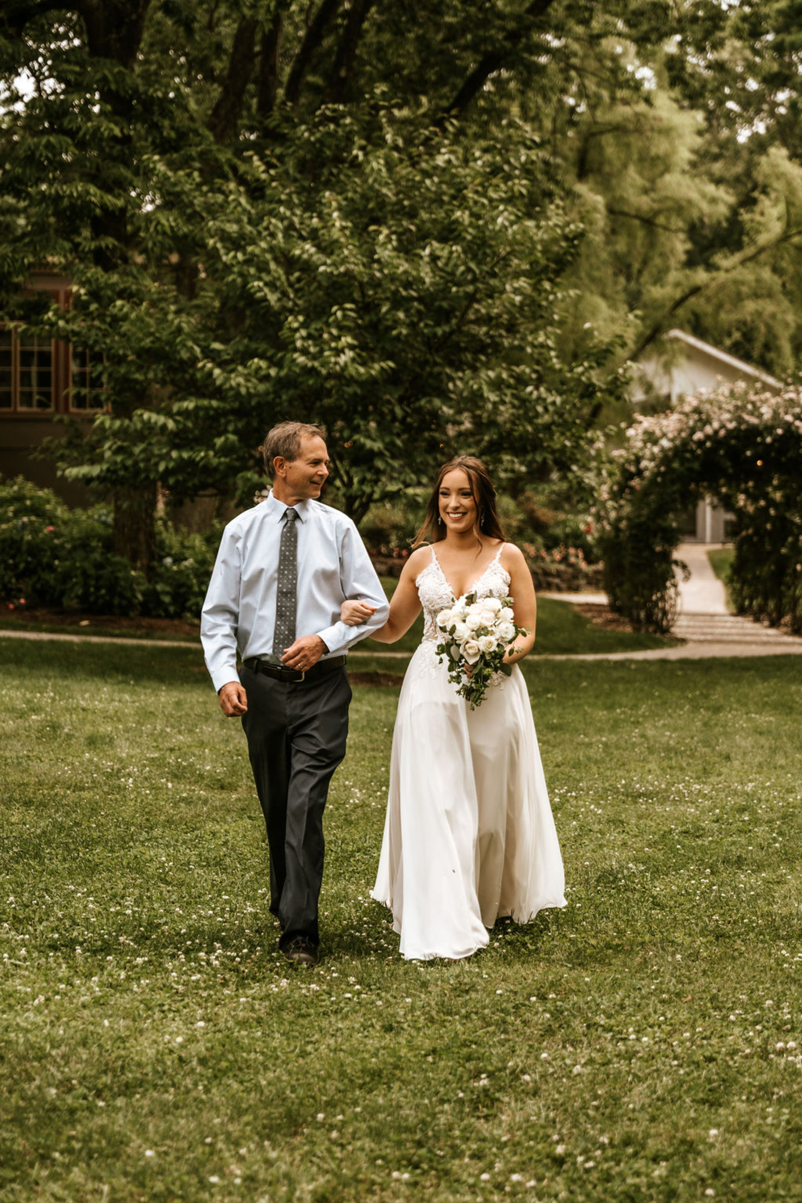 The bride walks arm in arm with her father through the gardens of Moonstone Manor, holding a white rose bouquet before the ceremony.