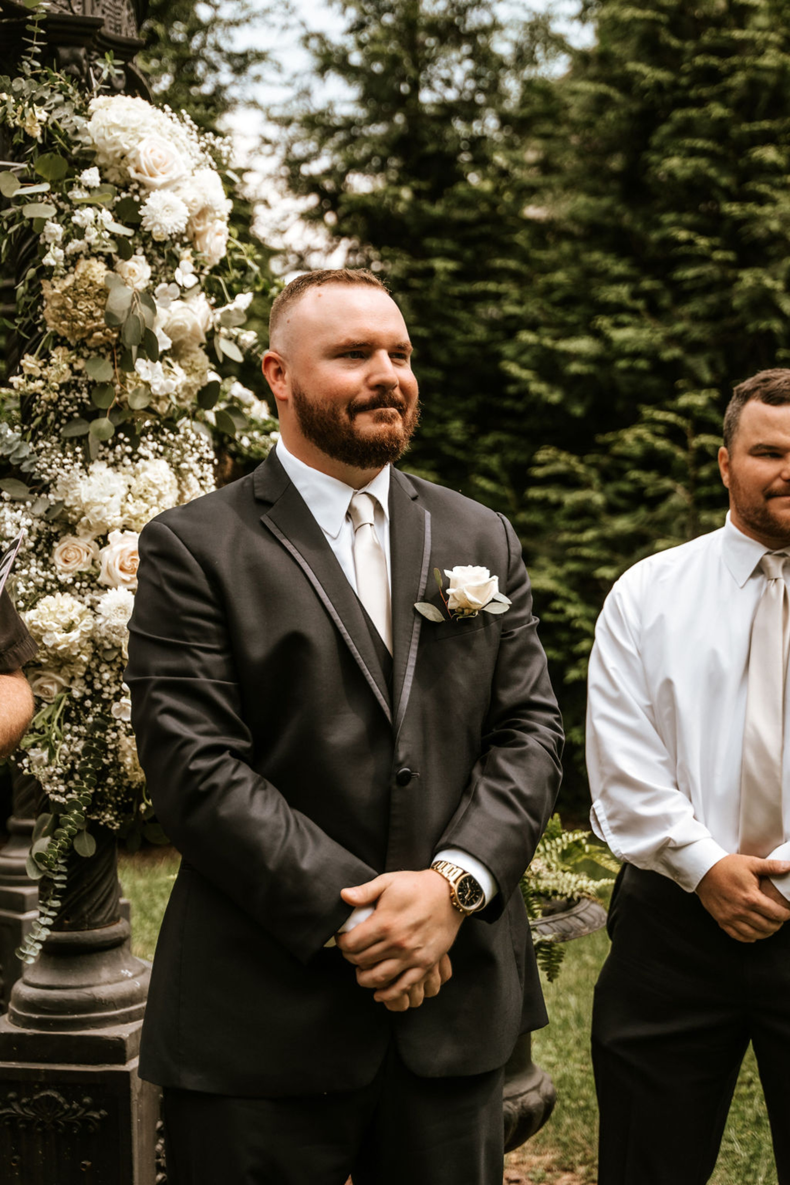 The groom stands smiling at the floral-adorned iron gazebo, waiting for his bride during their intimate Moonstone Manor wedding ceremony.