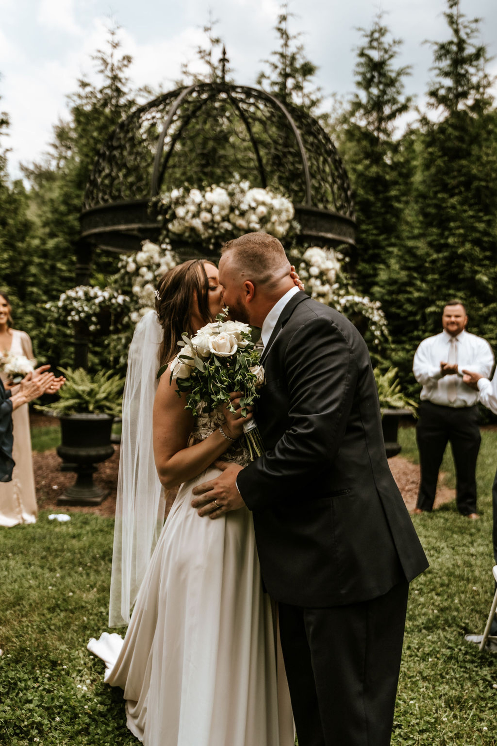The couple shares their first kiss beneath the floral-covered gazebo surrounded by greenery during their intimate Moonstone Manor ceremony.