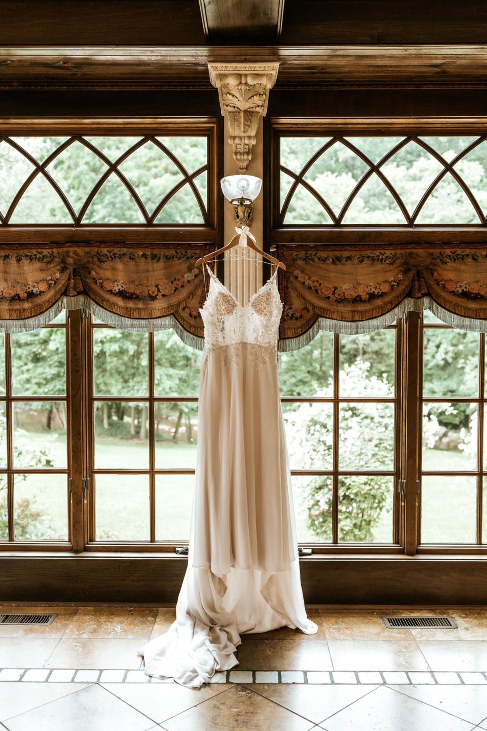 A romantic lace and chiffon wedding gown hangs gracefully in front of large windows inside Moonstone Manor, showcasing the venue’s historic charm and garden views.