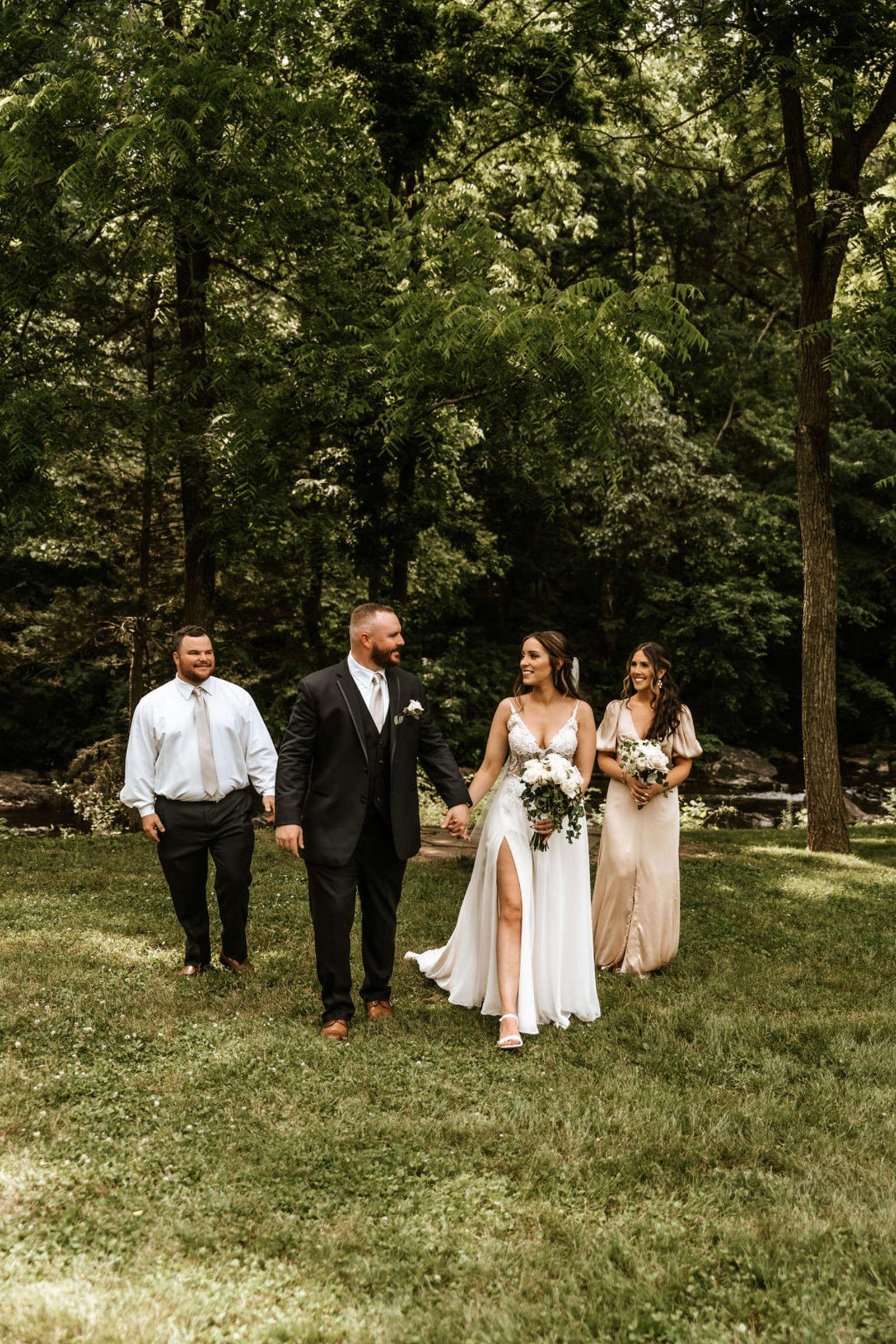 The bride and groom walk hand in hand with their wedding party through the wooded gardens at Moonstone Manor, celebrating their just-married moment.