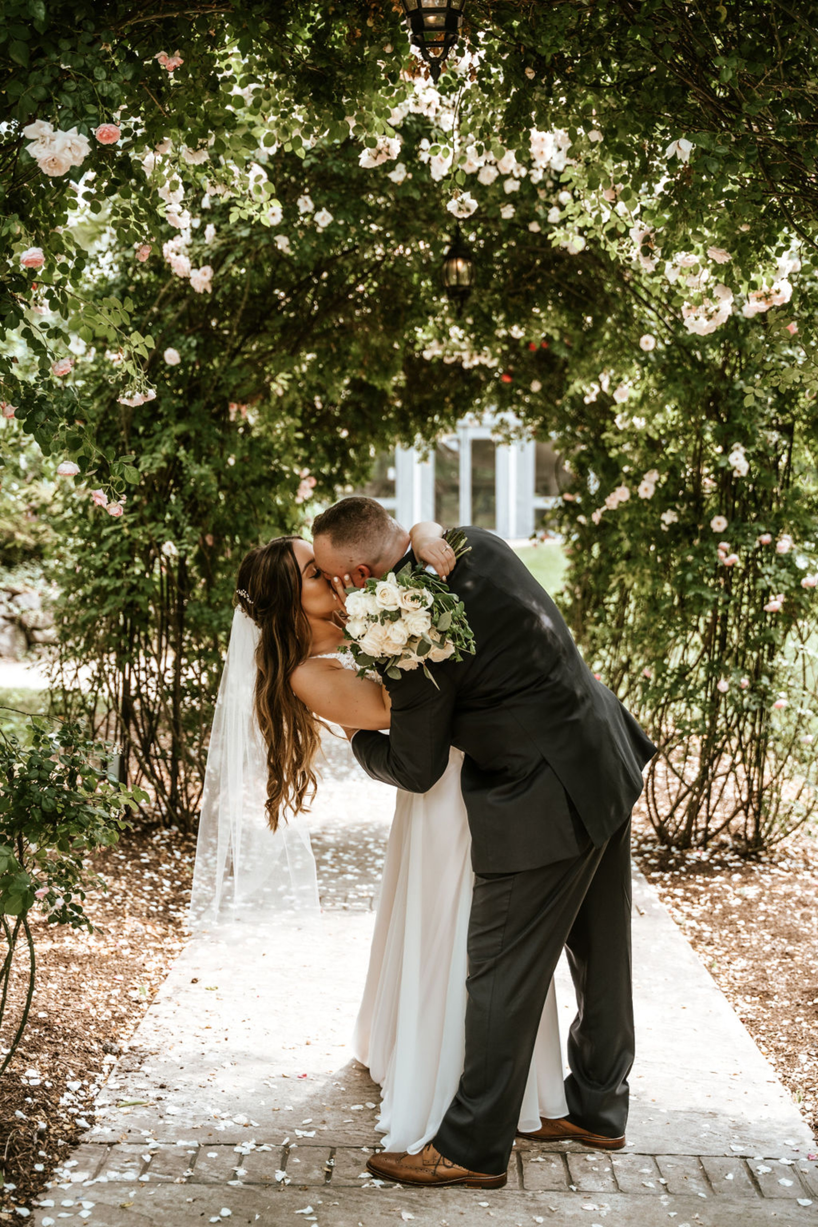 Bride and groom share a romantic kiss beneath a rose-covered archway at Moonstone Manor, surrounded by soft petals and lush summer greenery.