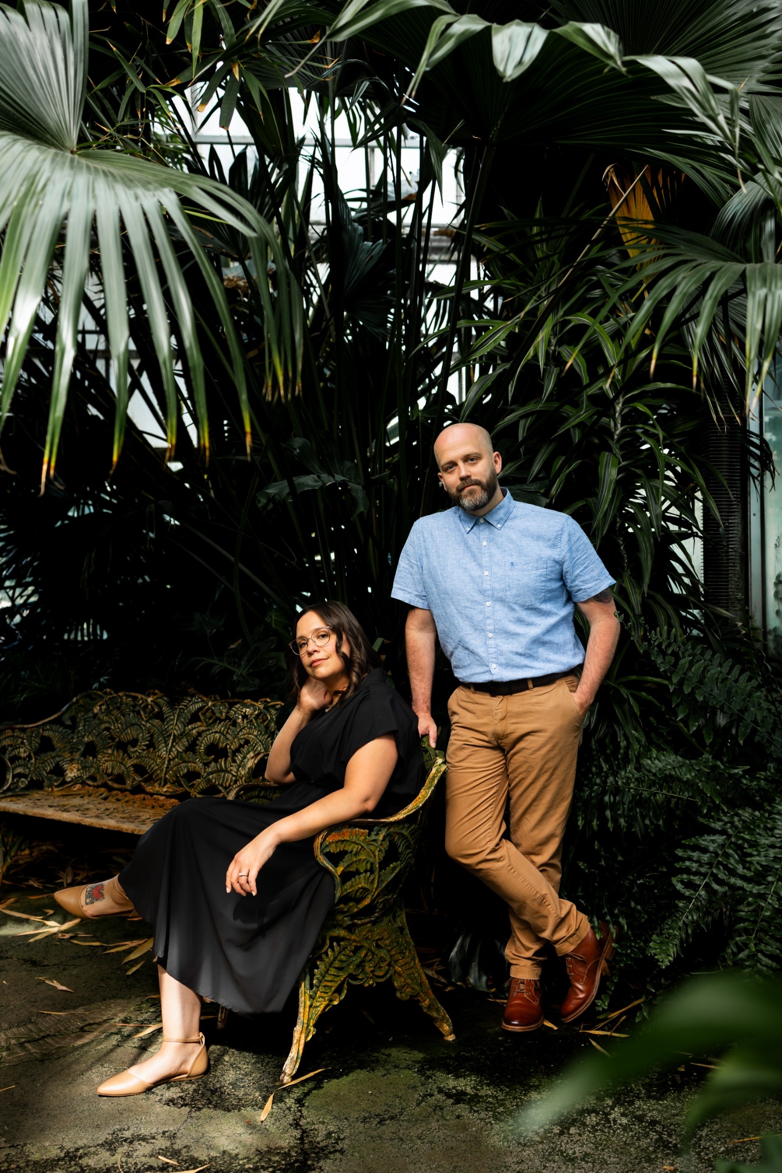 A couple poses for an engagement photo inside Ott’s Exotic Plants greenhouse, with the woman seated on a vintage bench and the man standing beside her, surrounded by tall palm leaves and rich greenery.