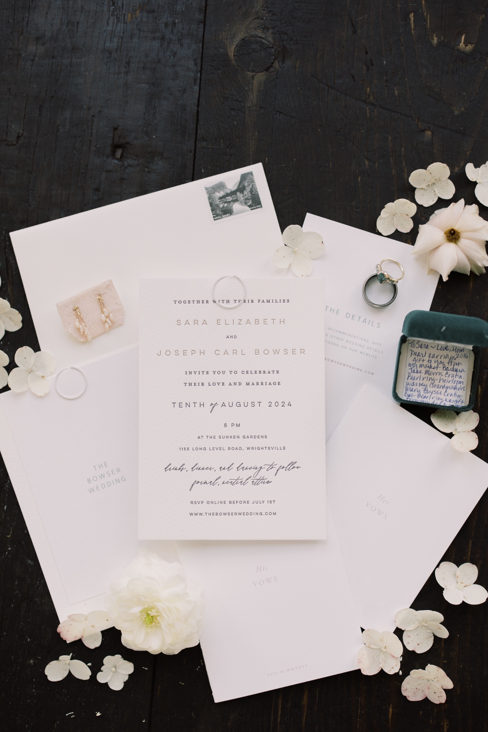 Invitation Suite Wedding Flat Lay with Vow Books and Rings Wedding flat lay with invitation suite, vow books, engagement ring, earrings, and white florals styled on a dark wood surface.