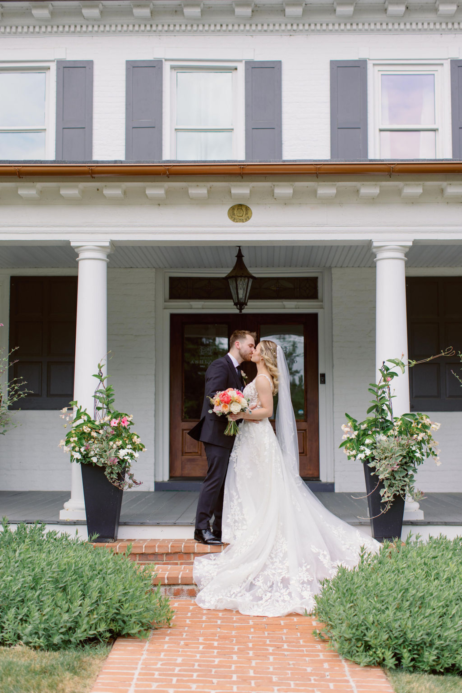 Bride and groom kissing on the front porch of Historic Ashland in Central Pennsylvania, surrounded by soft summer florals and lush greenery during their romantic early summer wedding.