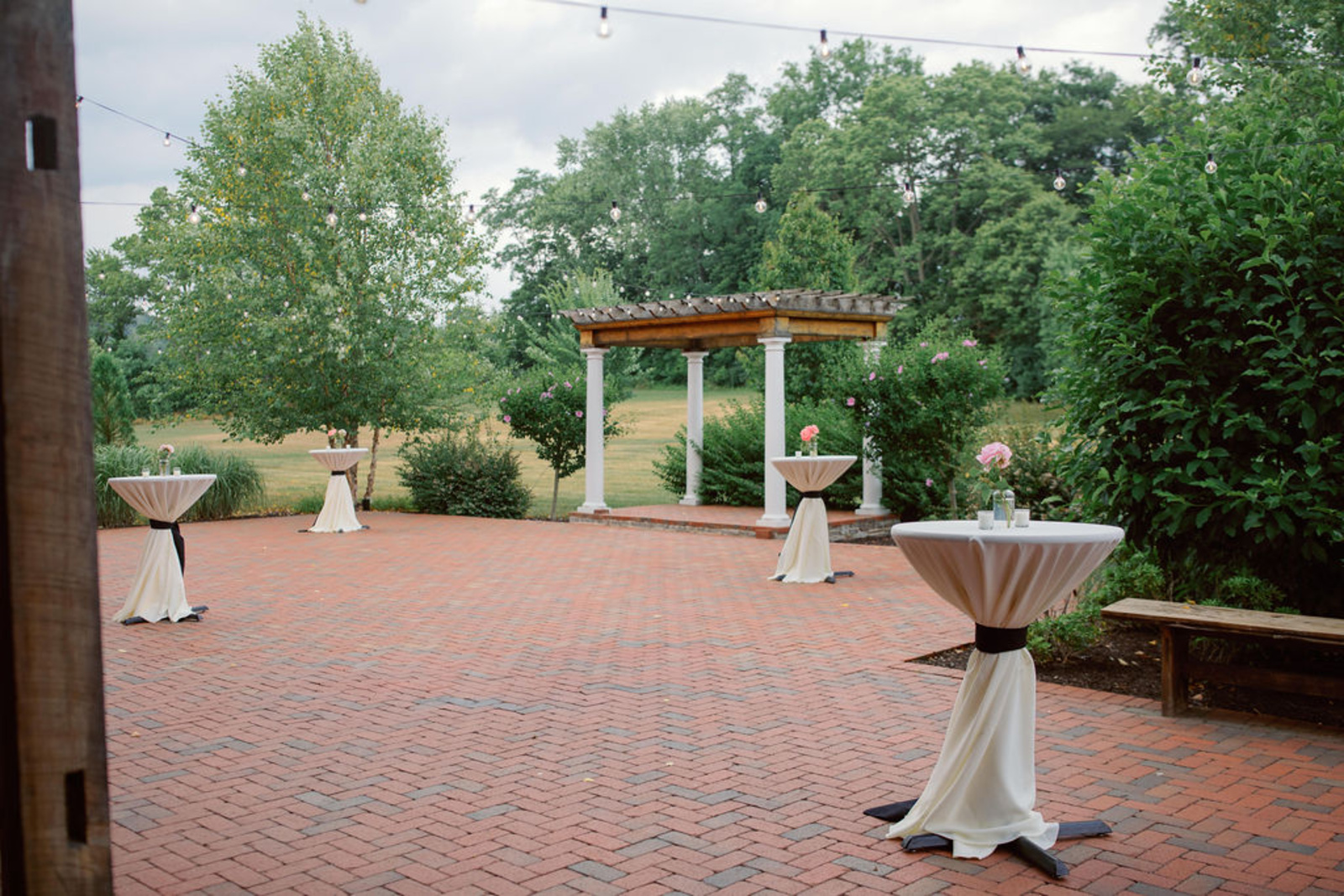 Brick courtyard cocktail hour space at Historic Ashland featuring string lights, greenery, and cocktail tables before a summer wedding reception.