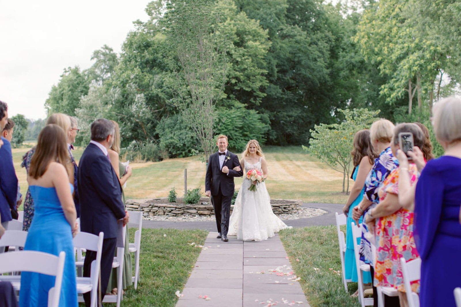 Bride walking down the outdoor ceremony aisle with a family member during her early summer wedding at Historic Ashland in Pennsylvania.