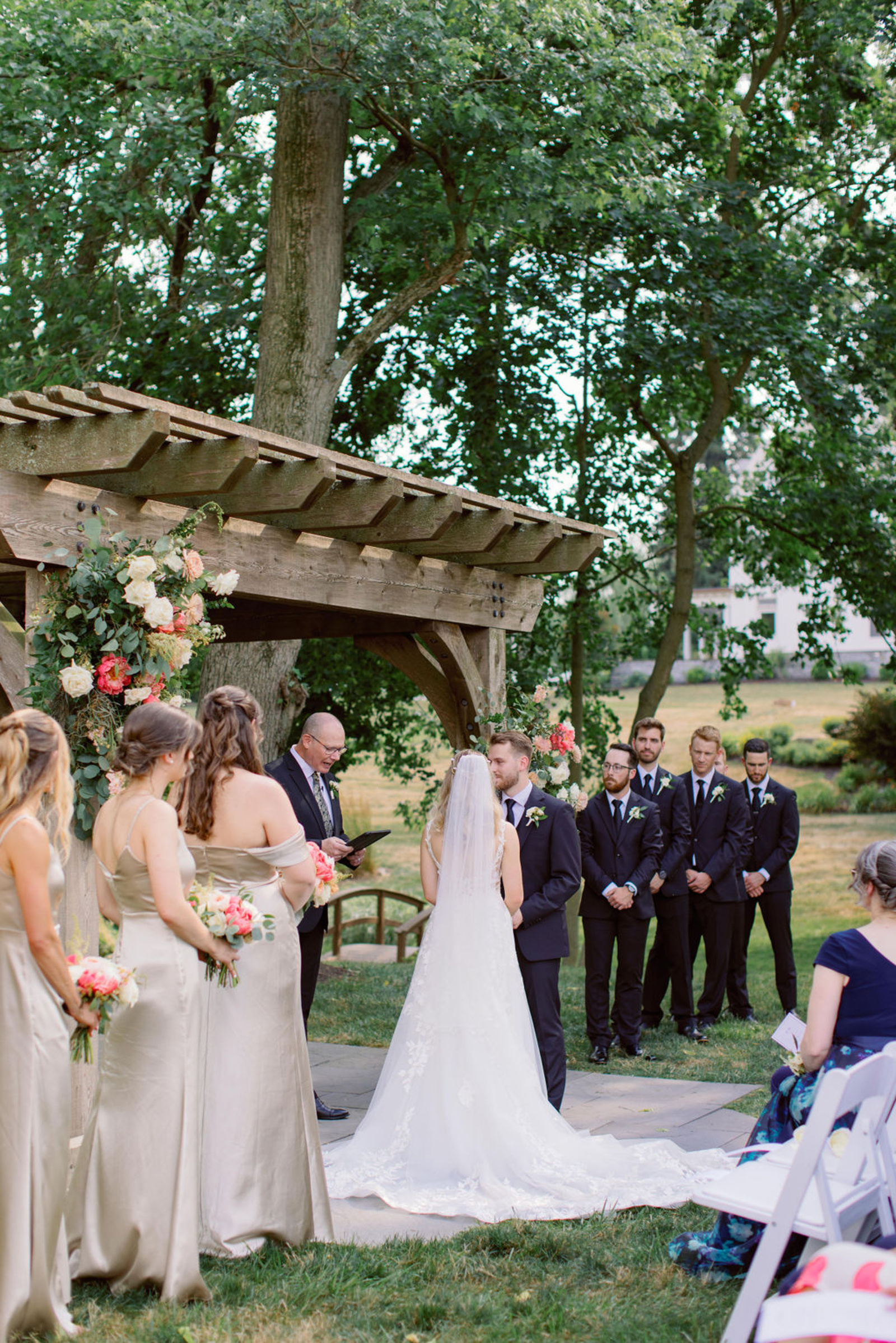 Bride and groom exchanging vows beneath a wooden pergola decorated with summer florals during an outdoor wedding ceremony at Historic Ashland.