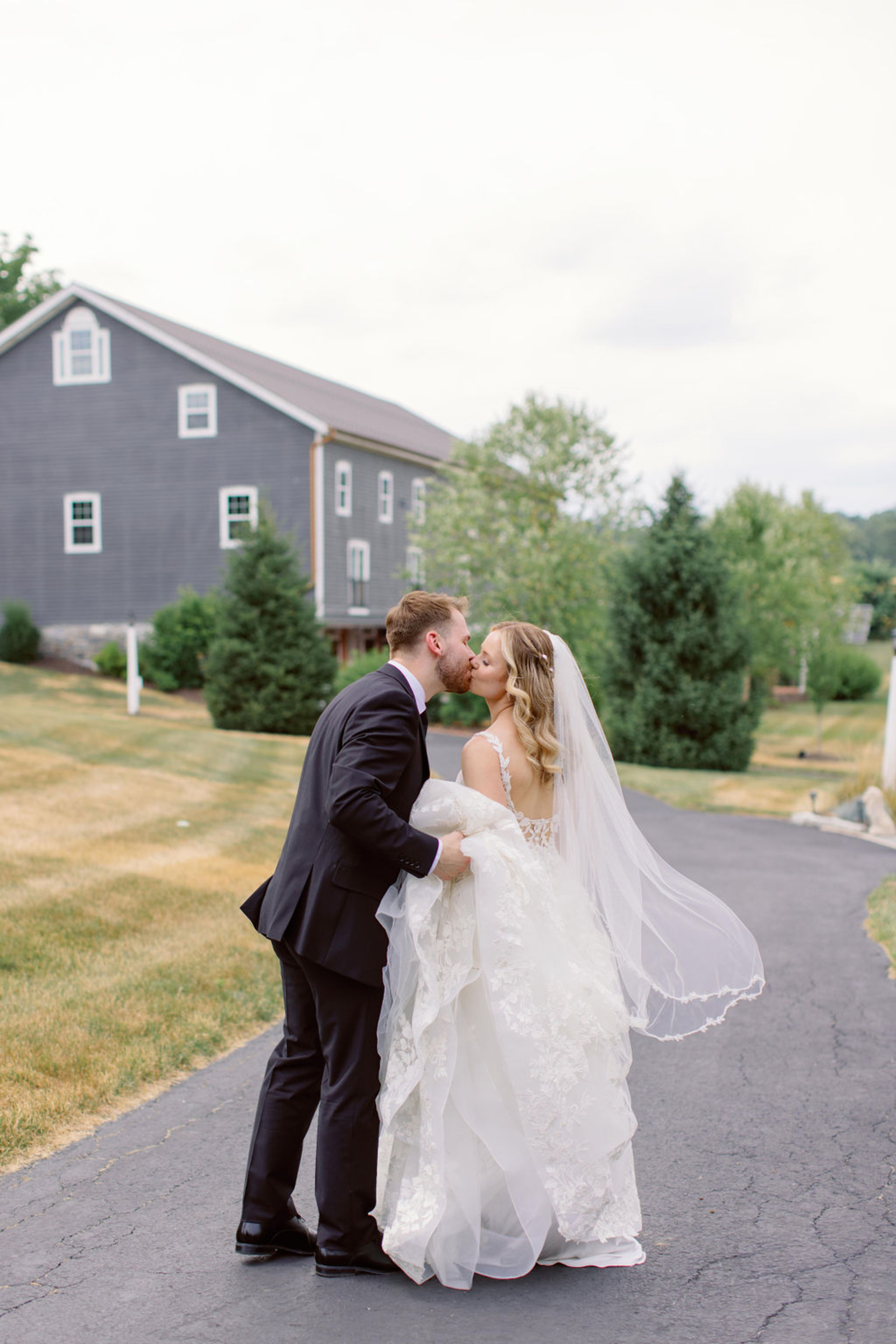 Bride and groom kissing on the driveway outside Historic Ashland with the reception barn in the background during their summer wedding portraits.