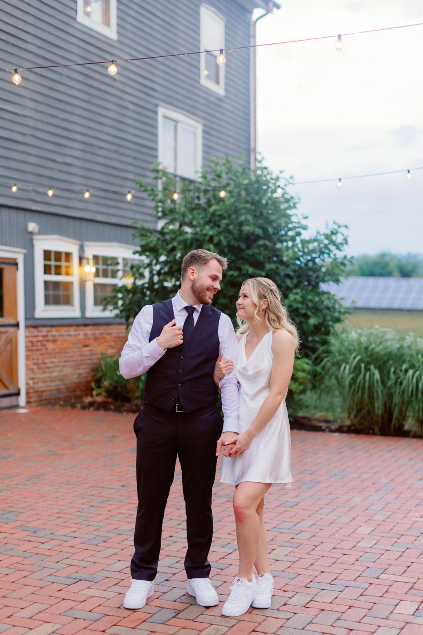 Bride and groom in reception outfits walking hand-in-hand outside Historic Ashland under string lights at dusk.