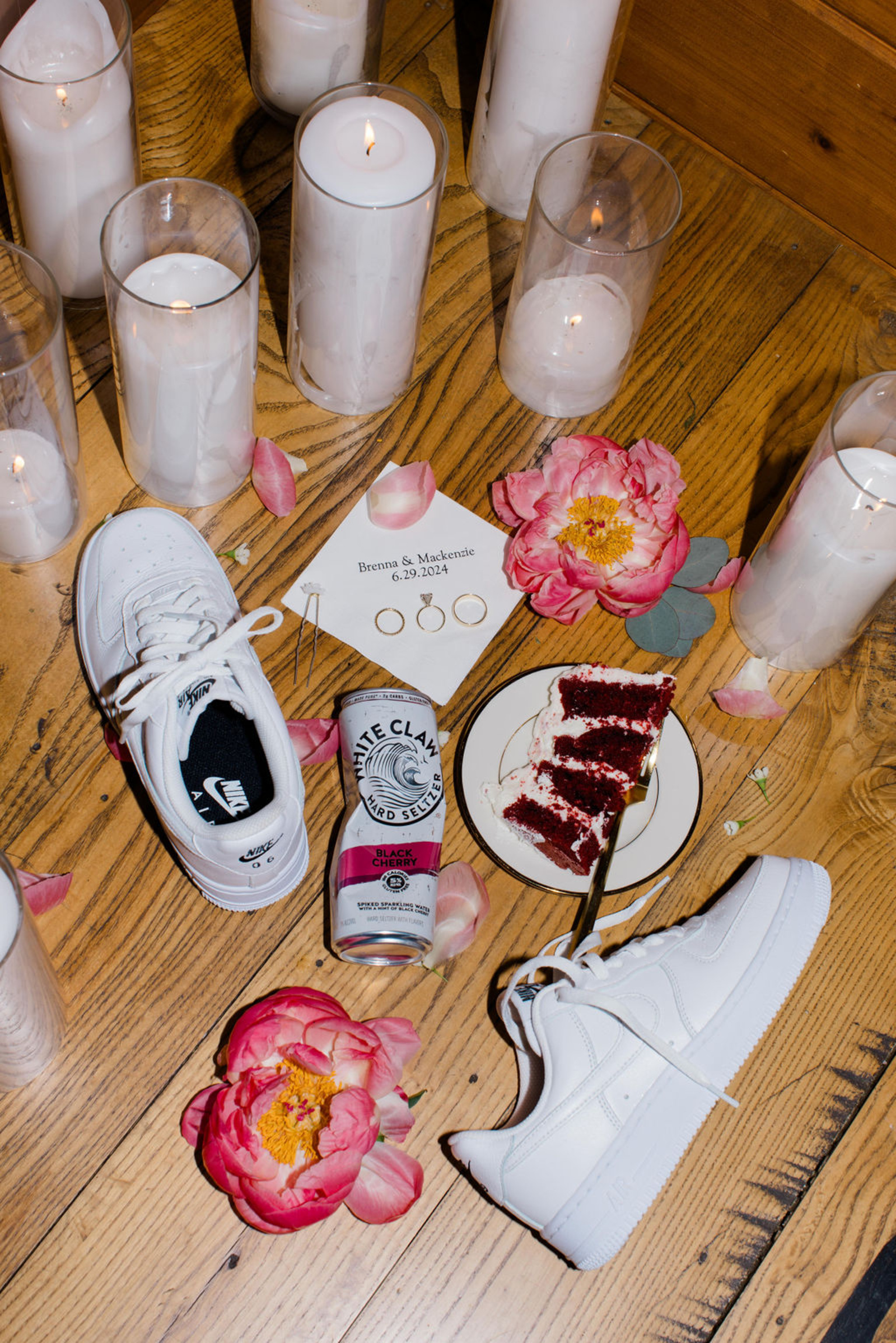 End of the night wedding flat lay featuring white Nike sneakers, pink florals, wedding rings, candles, cake, and a White Claw on a wood floor.
