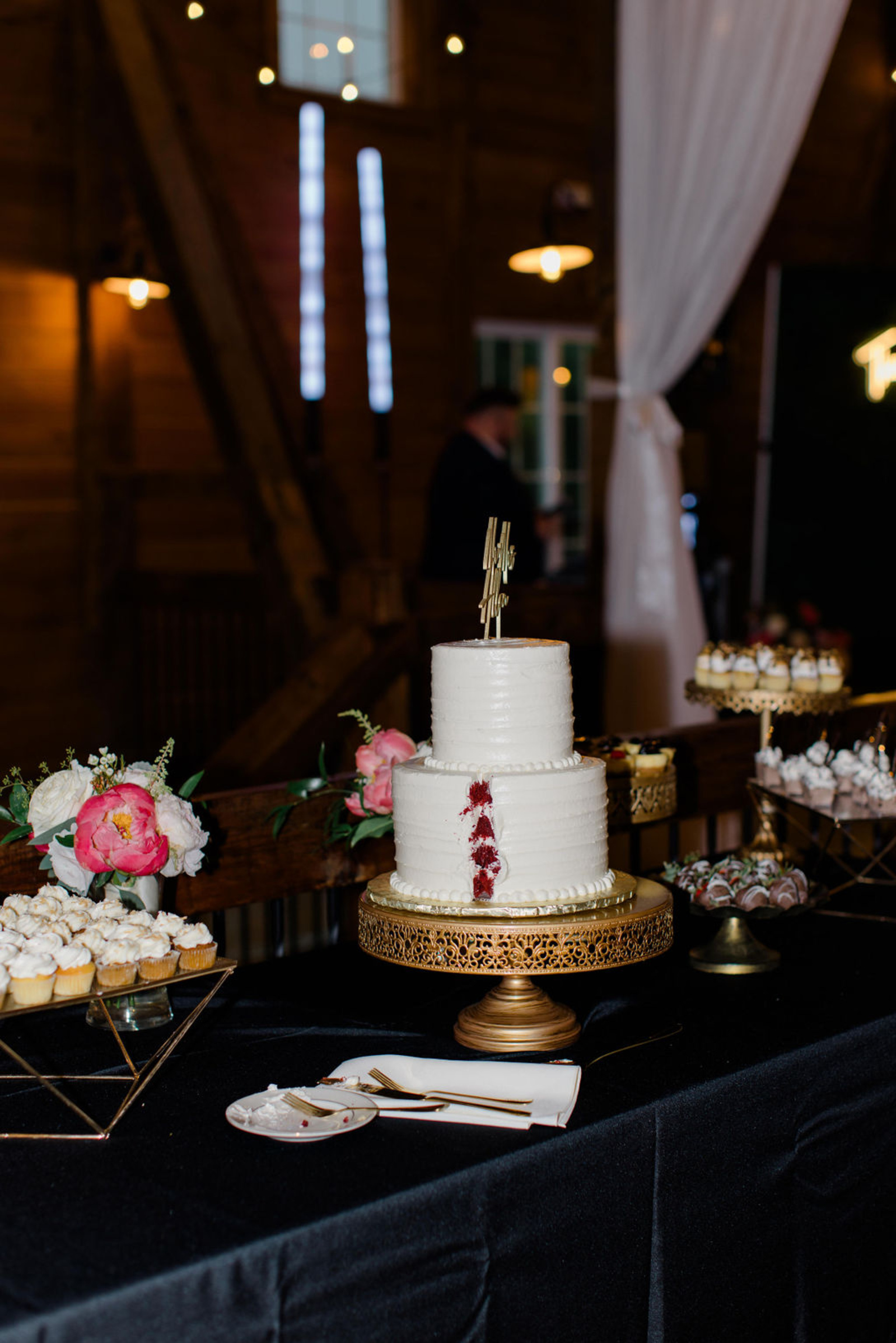 White wedding cake with pink florals and desserts displayed inside Historic Ashland’s candlelit barn reception space.