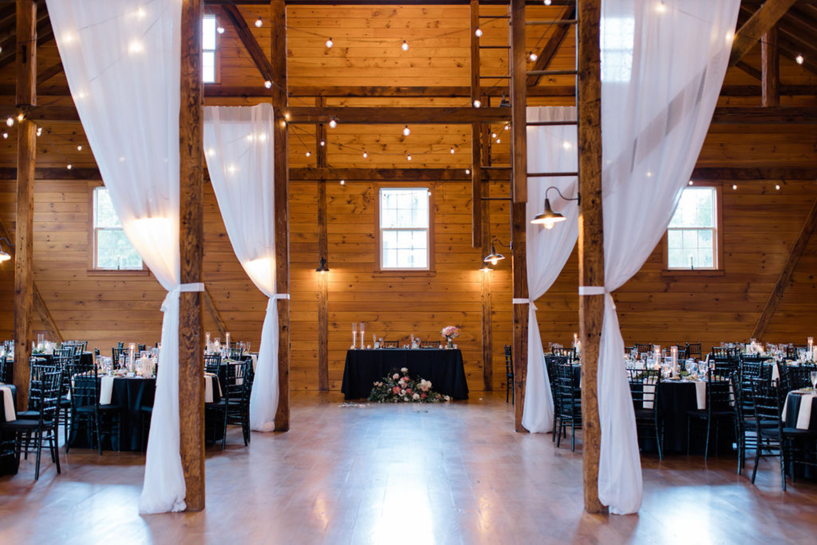 Romantic barn reception space at Historic Ashland with wood beams, draped white fabric, string lights, and black-and-white reception tables.