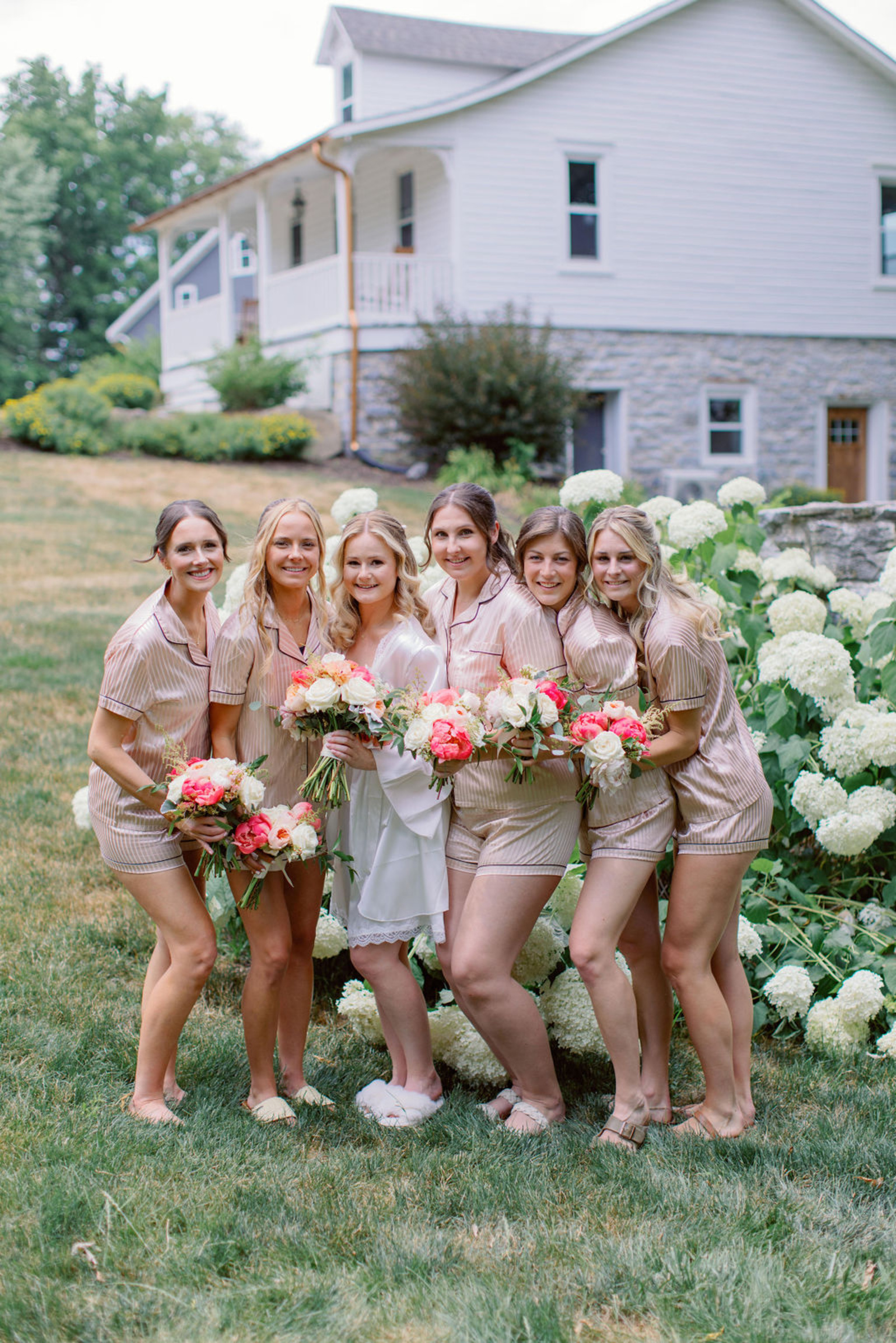 Bride and bridesmaids in matching champagne-toned pajamas holding colorful summer bouquets outside Historic Ashland before the wedding ceremony.