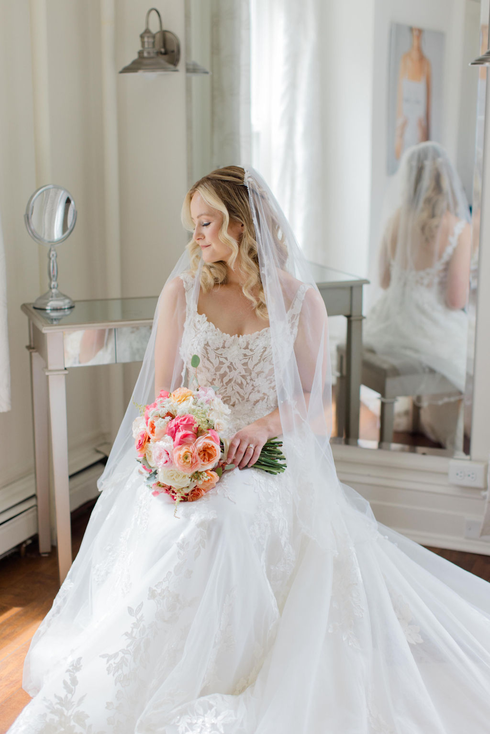 Bride sitting in her lace wedding gown holding a coral and blush bouquet inside Historic Ashland during getting-ready moments before the ceremony.