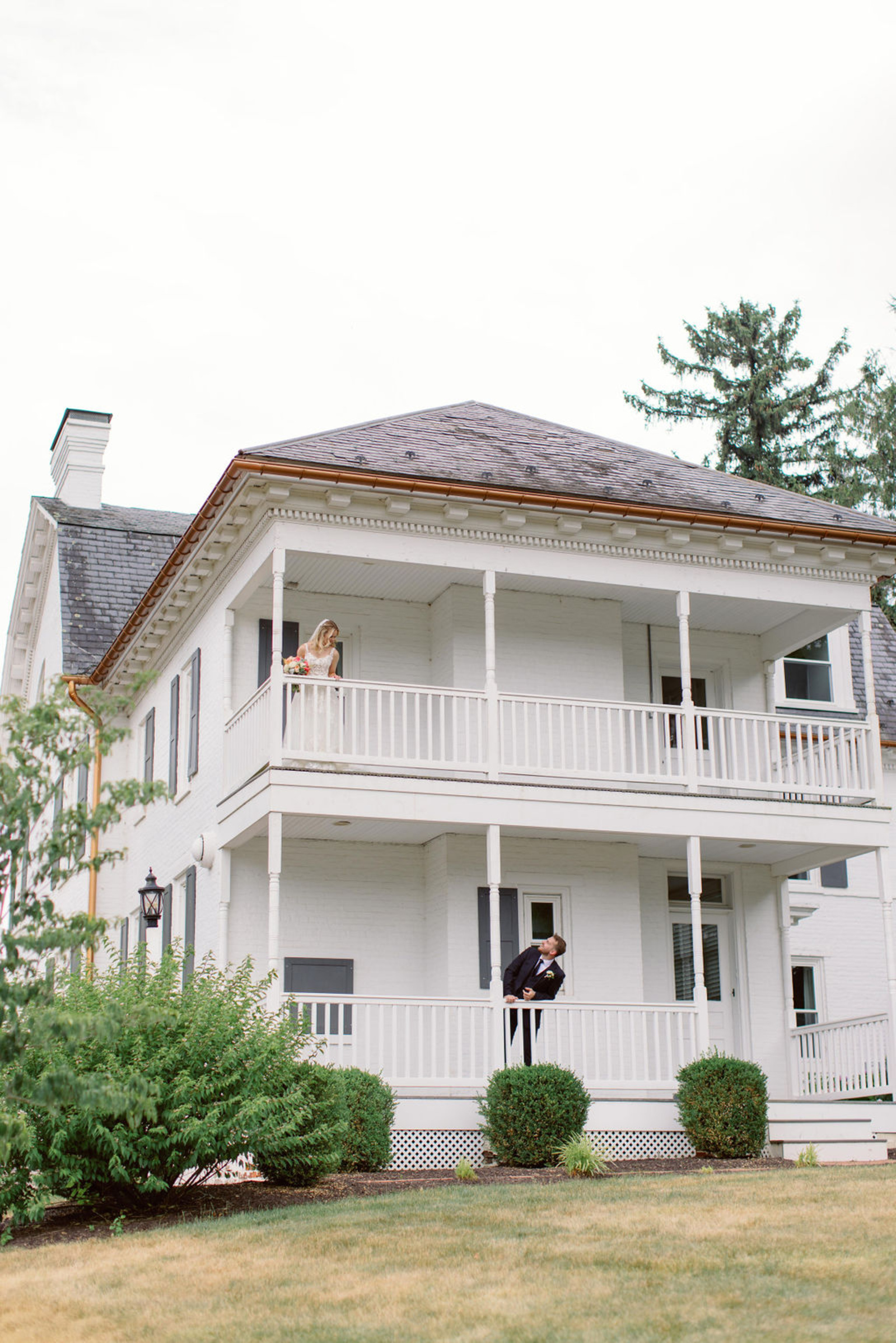 Bride standing on the second-story balcony while the groom looks up from the porch below at Historic Ashland during their early summer Pennsylvania wedding.