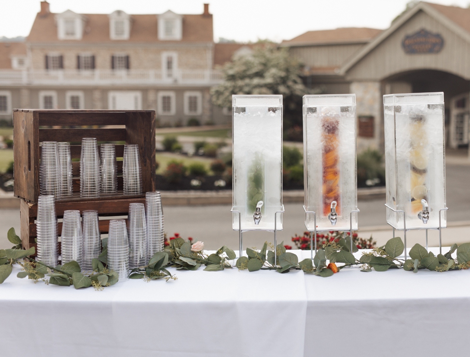 Outdoor wedding beverage station with three clear drink dispensers filled with fruit-infused water and stacked plastic cups in a wooden crate, decorated with greenery and floral accents on a white tablecloth.