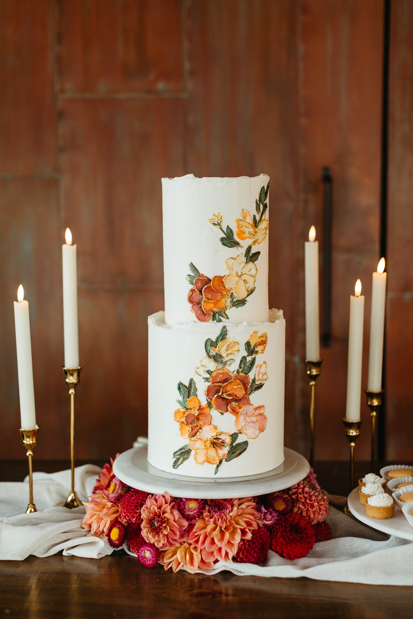 Colorful Floral Wedding Cake Two-tier white wedding cake with painted orange and peach flowers surrounded by candles and dahlias.