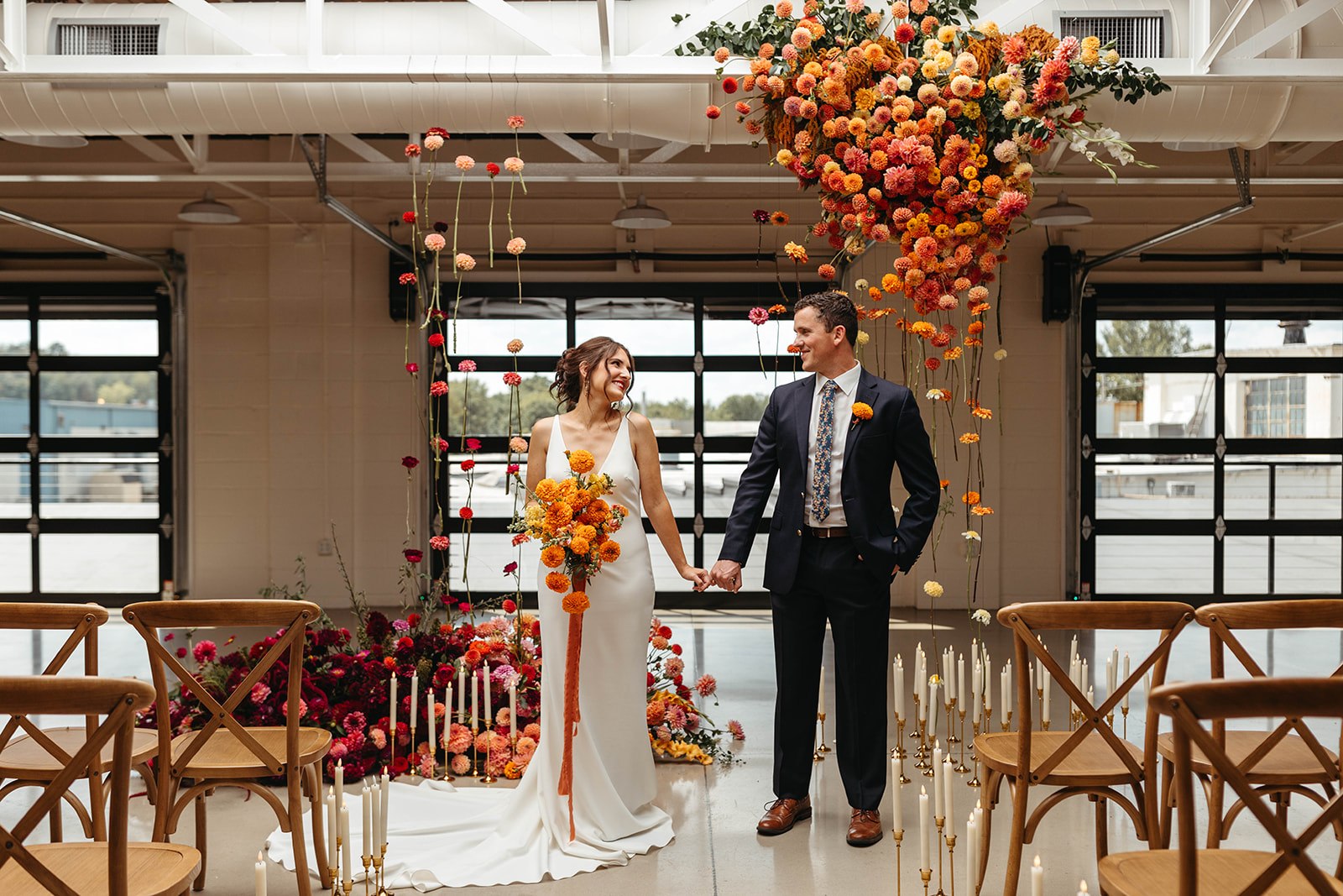 Colorful Industrial Wedding at The Booking House in Manheim, PA Bride and groom standing beneath a colorful hanging floral installation inside The Booking House in Manheim, PA, surrounded by candles and industrial-style garage doors.