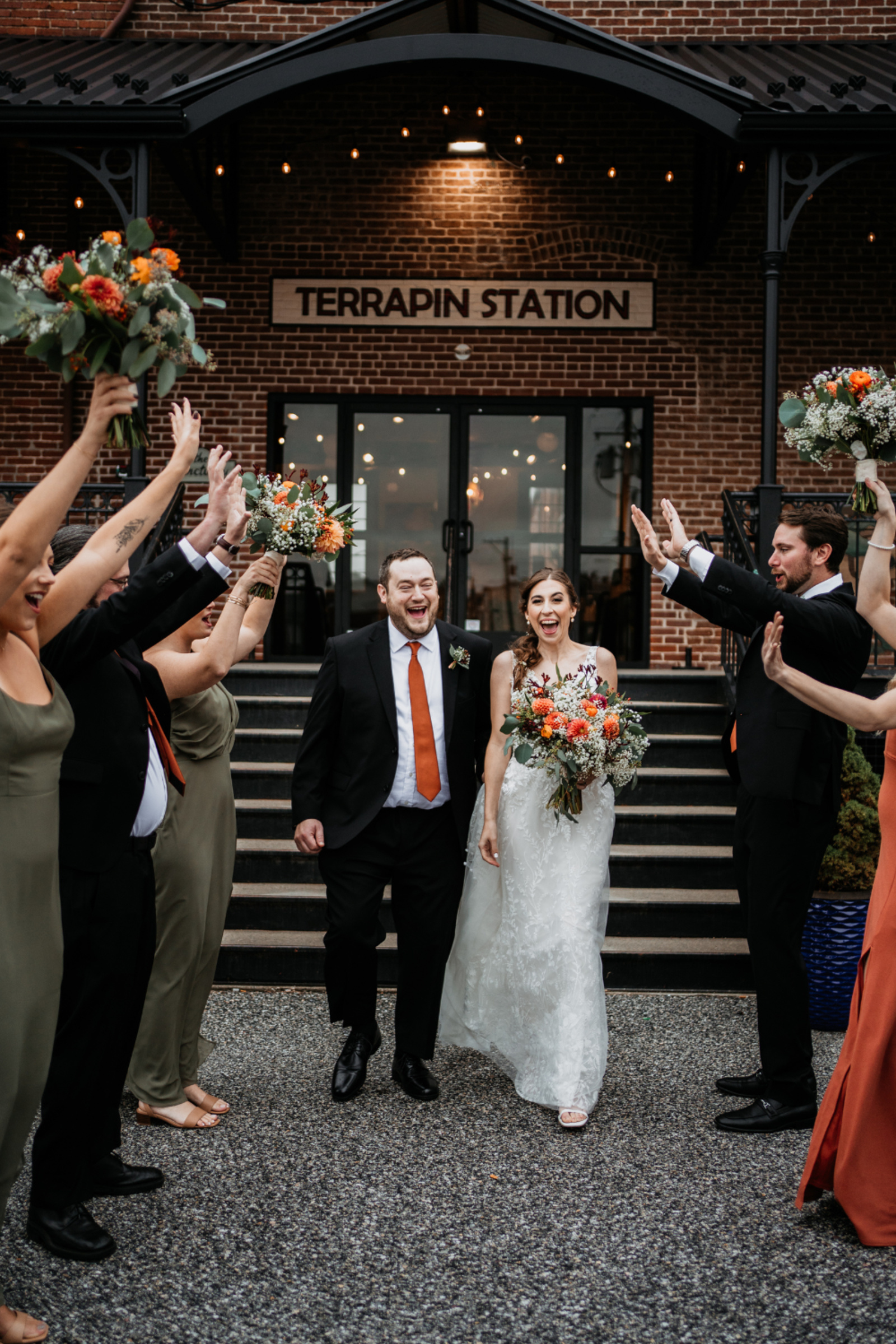 Newlyweds walk through cheering bridal party outside Terrapin Station, holding colorful bouquets and smiling with excitement.
