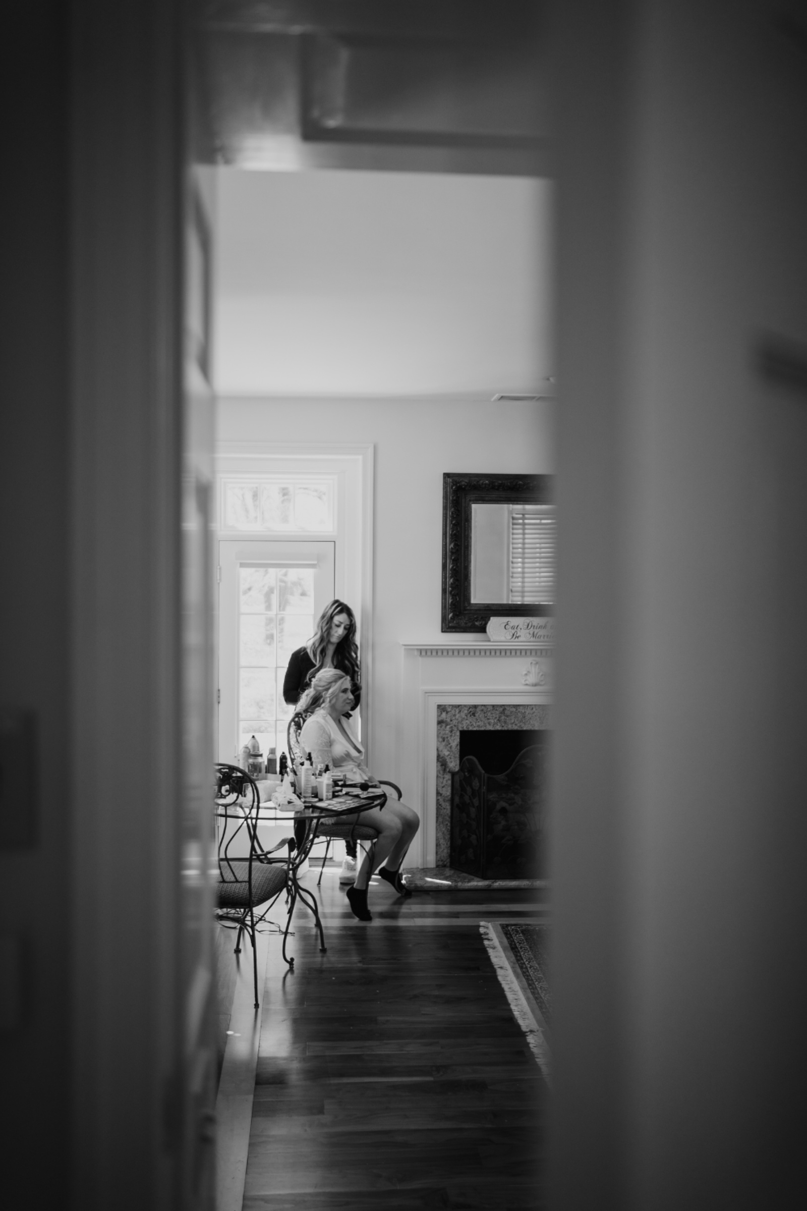 Black and white photo taken through a doorway showing a bride sitting by a window as a stylist works on her hair, capturing a quiet wedding morning moment.