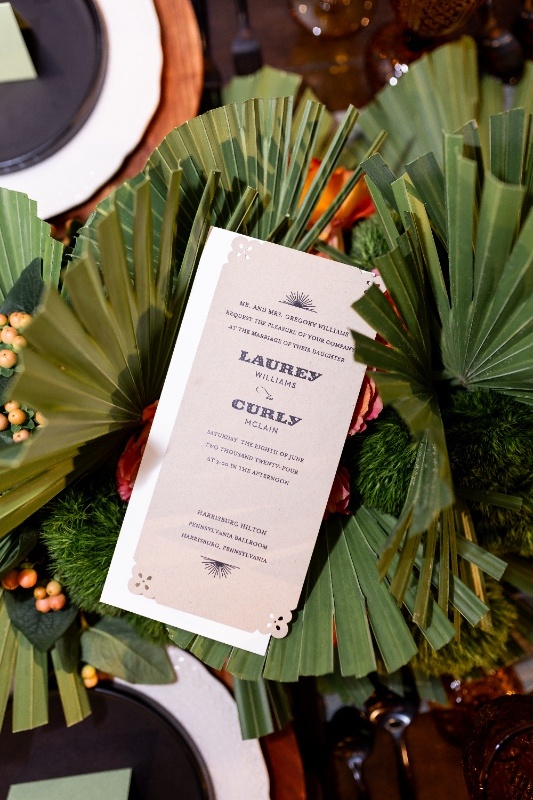 Oklahoma!-Inspired Wedding Invitation with Rustic Western Flair A rustic wedding invitation featuring the names Laurey and Curly rests among green palm leaves, orange roses, and textured greenery in a western-inspired Oklahoma! styled shoot.