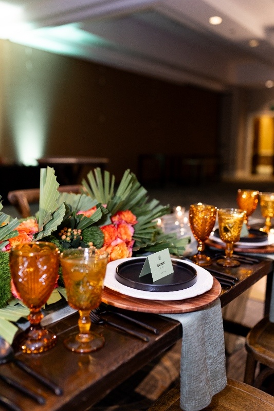 Bold Western-Inspired Wedding Table Setting A rustic wedding tablescape with amber glassware, black flatware, and layered plates featuring bold floral centerpieces of orange roses and fan-shaped greenery, inspired by Oklahoma!.