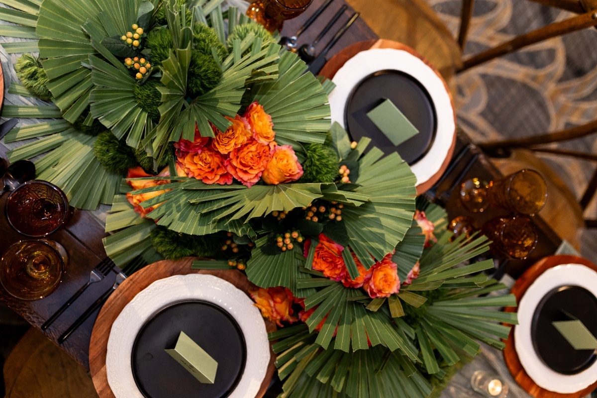 Vibrant Floral Centerpiece with Western Charm A top-down view of a wedding table featuring lush greenery, palm fans, and bright orange roses arranged in a bold Oklahoma!-inspired centerpiece.