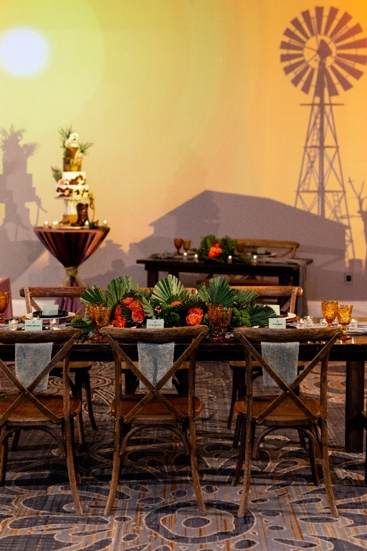 Western-Inspired Reception Setup with Warm Sunset Tones A rustic wedding reception table with amber glasses, greenery, and bold orange florals is set beneath warm lighting and a backdrop featuring a windmill silhouette.