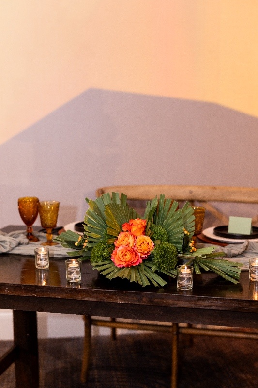 Rustic Wedding Centerpiece with Orange Roses and Palm Leaves A wedding centerpiece made of green palms, orange roses, and soft candlelight adds western elegance to a rustic sweetheart table